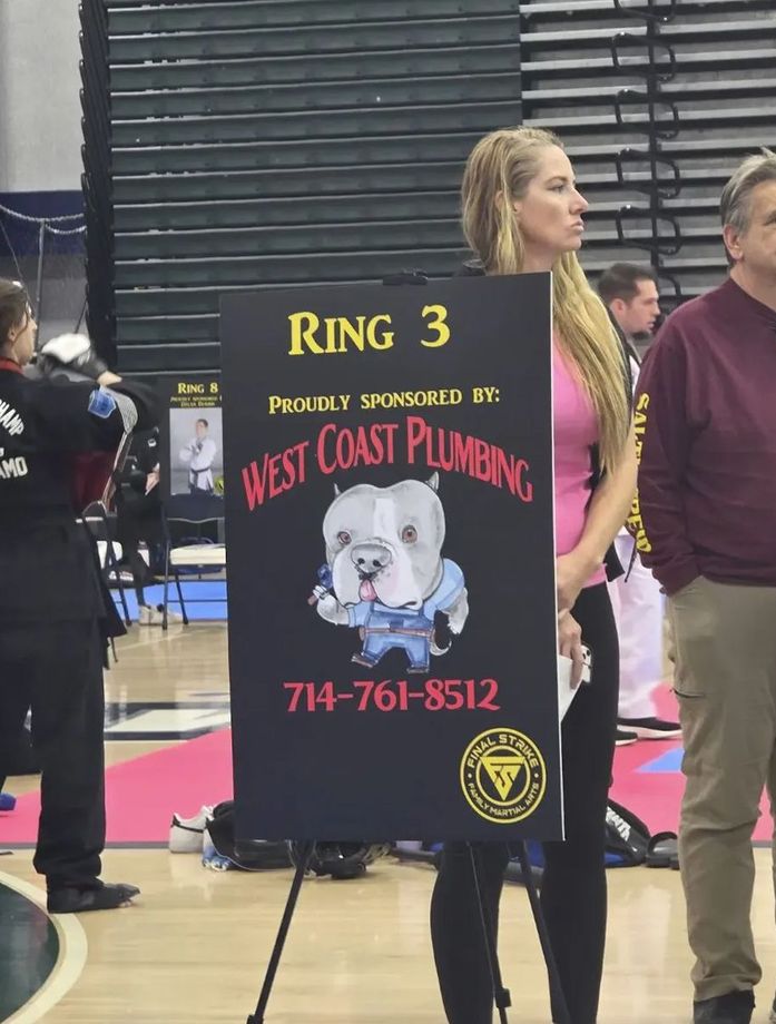Woman holding Ring 3 sign for West Coast Plumbing at an indoor event, with purple carpet and black blinds behind.