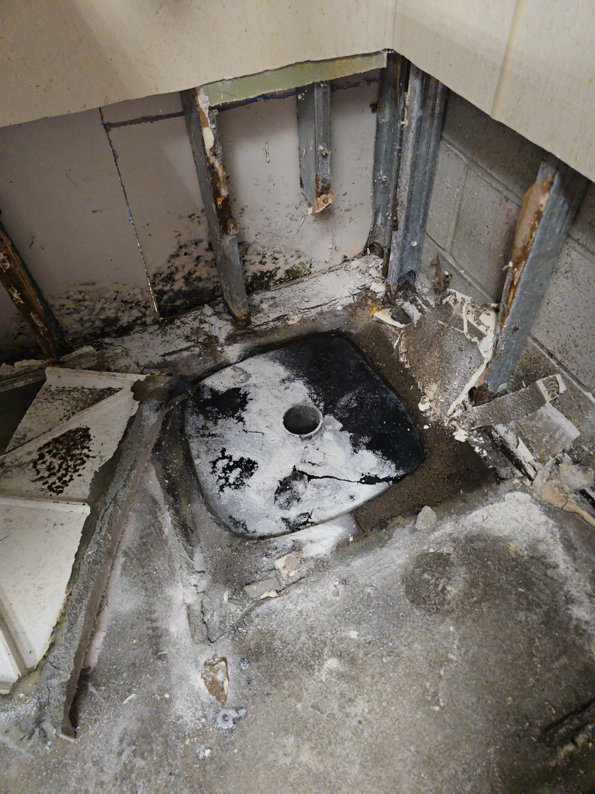 Damaged bathroom floor with exposed plumbing and a black-stained sink area, showing water damage and grime