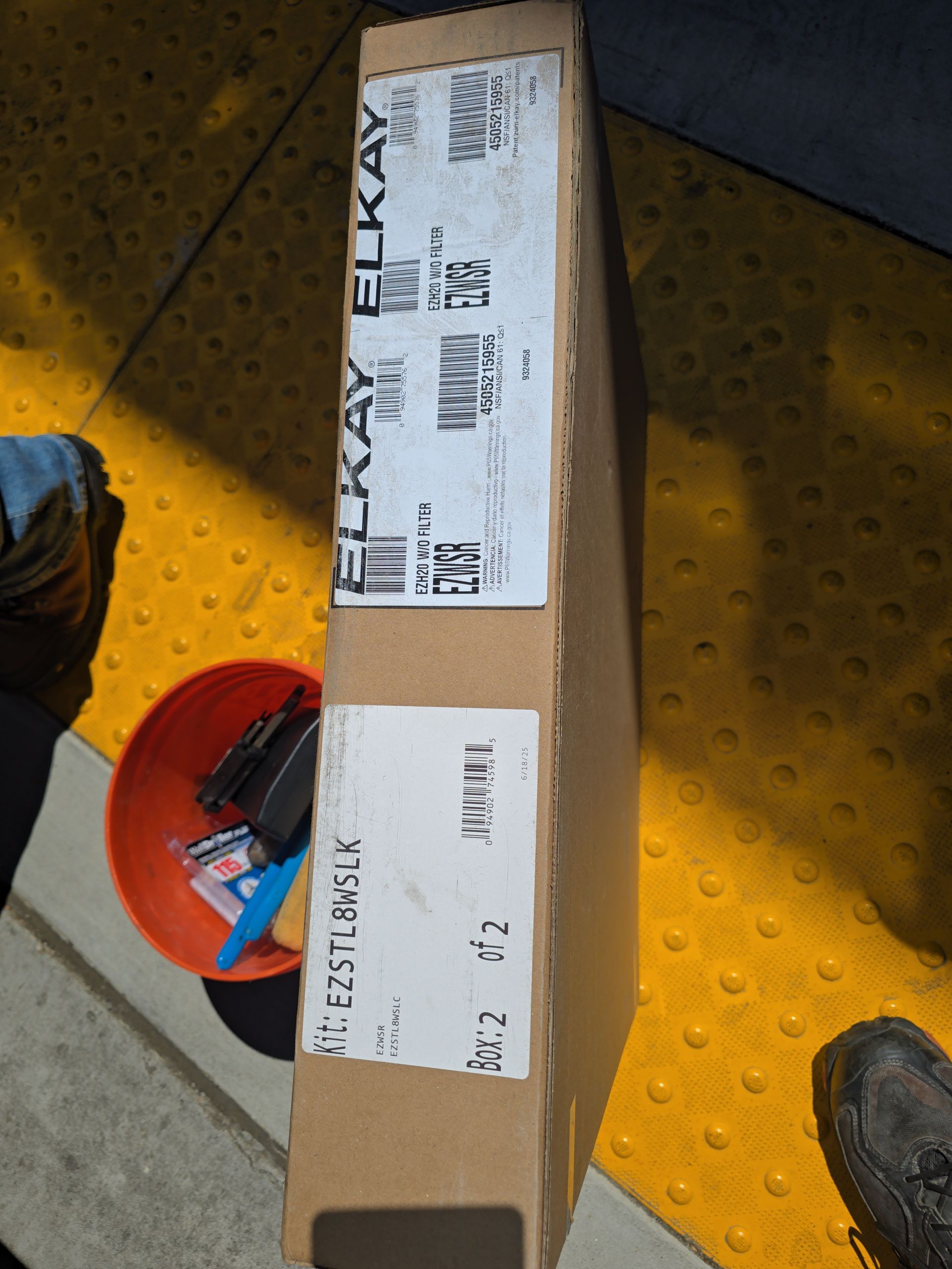Cardboard shipping box with white labels on a concrete floor beside a red tool, near two shoes.