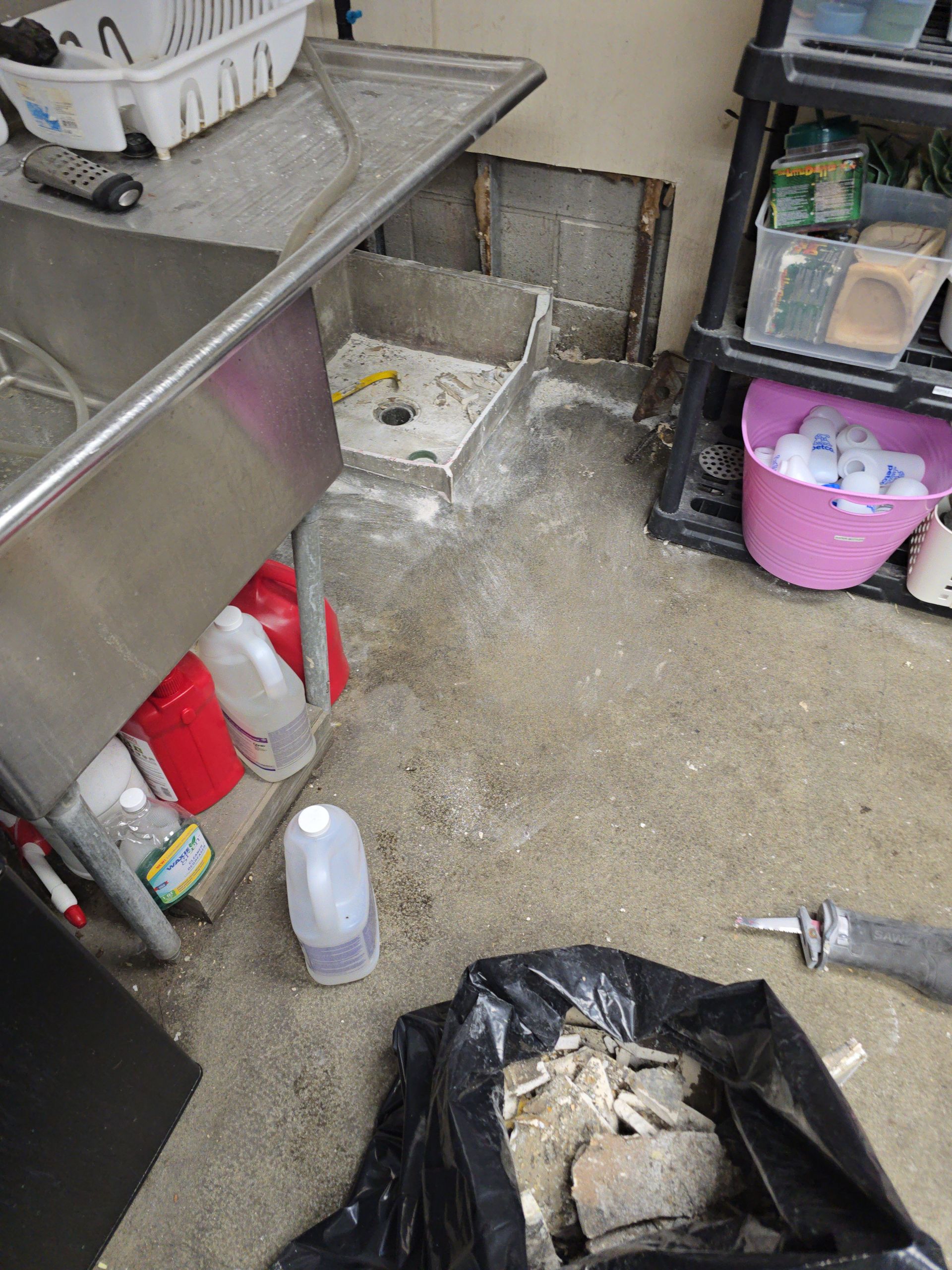 Dirty kitchen floor beside sink, cabinet, and cleaning supplies with a black trash bag in the foreground