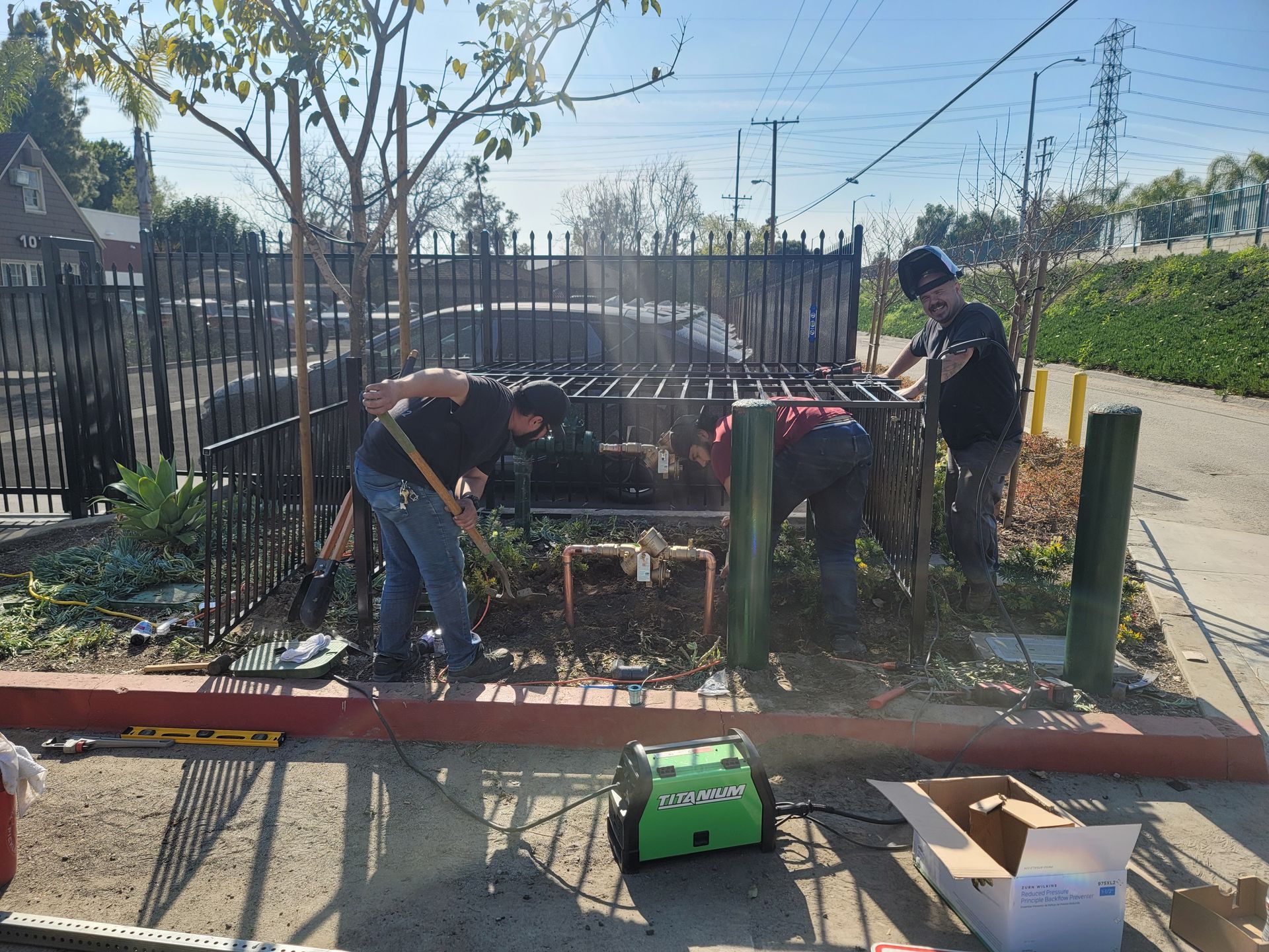 Workers fixing a fence in a sunny yard, with tools and materials on the ground.