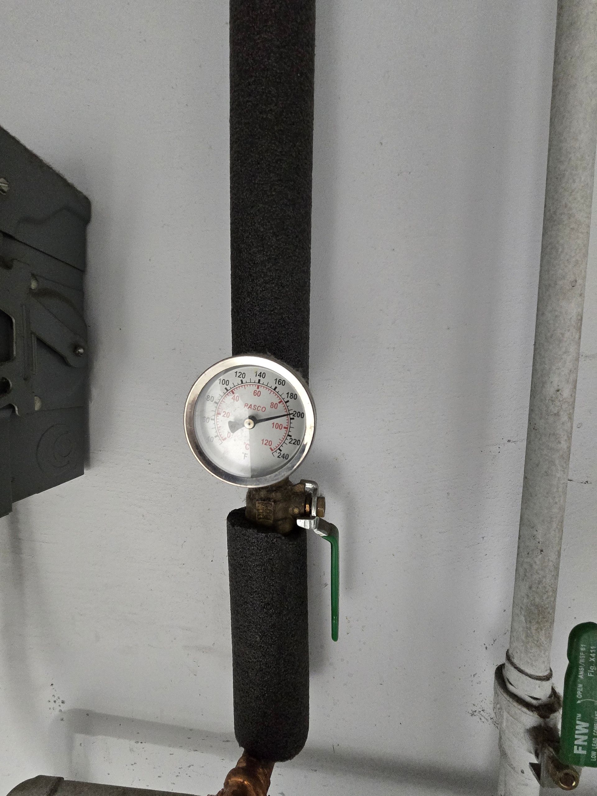 Pressure gauge on a pipe with black insulation and green valve handle against a gray wall.