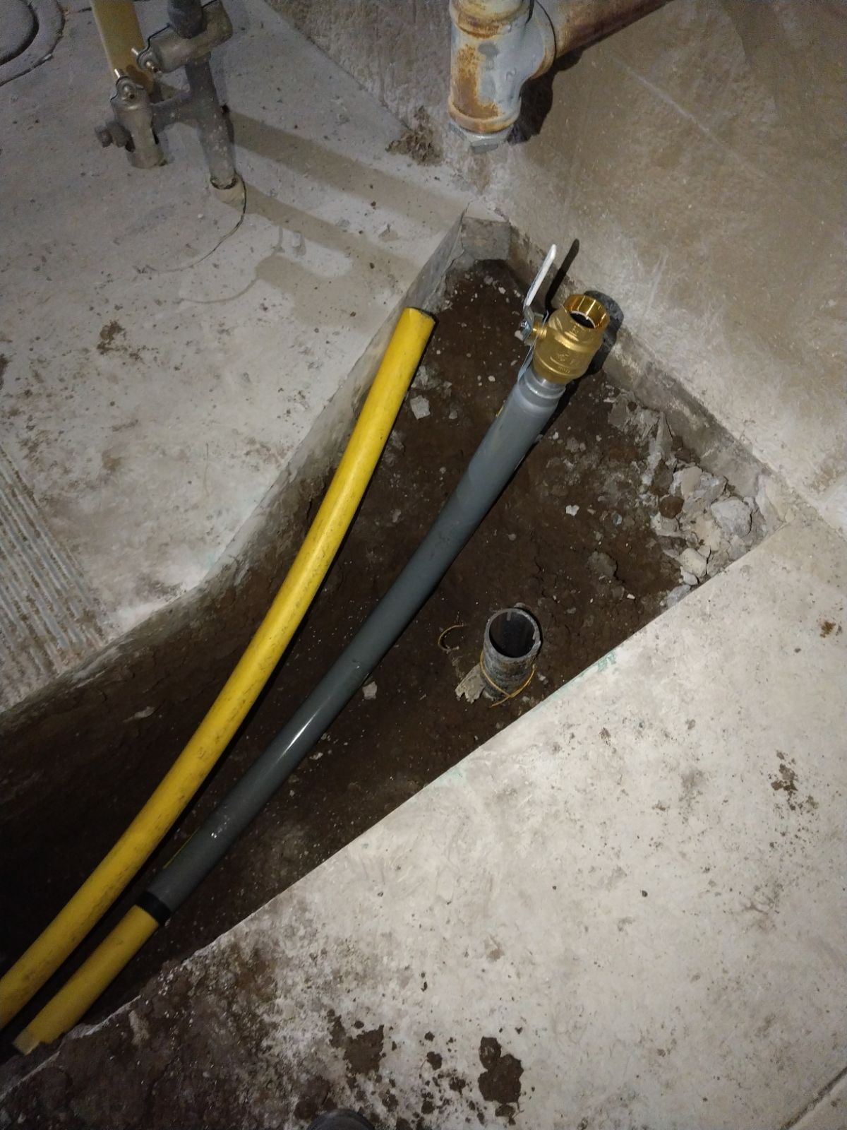 Concrete utility trench with yellow and gray hoses and a small pipe opening in a basement floor