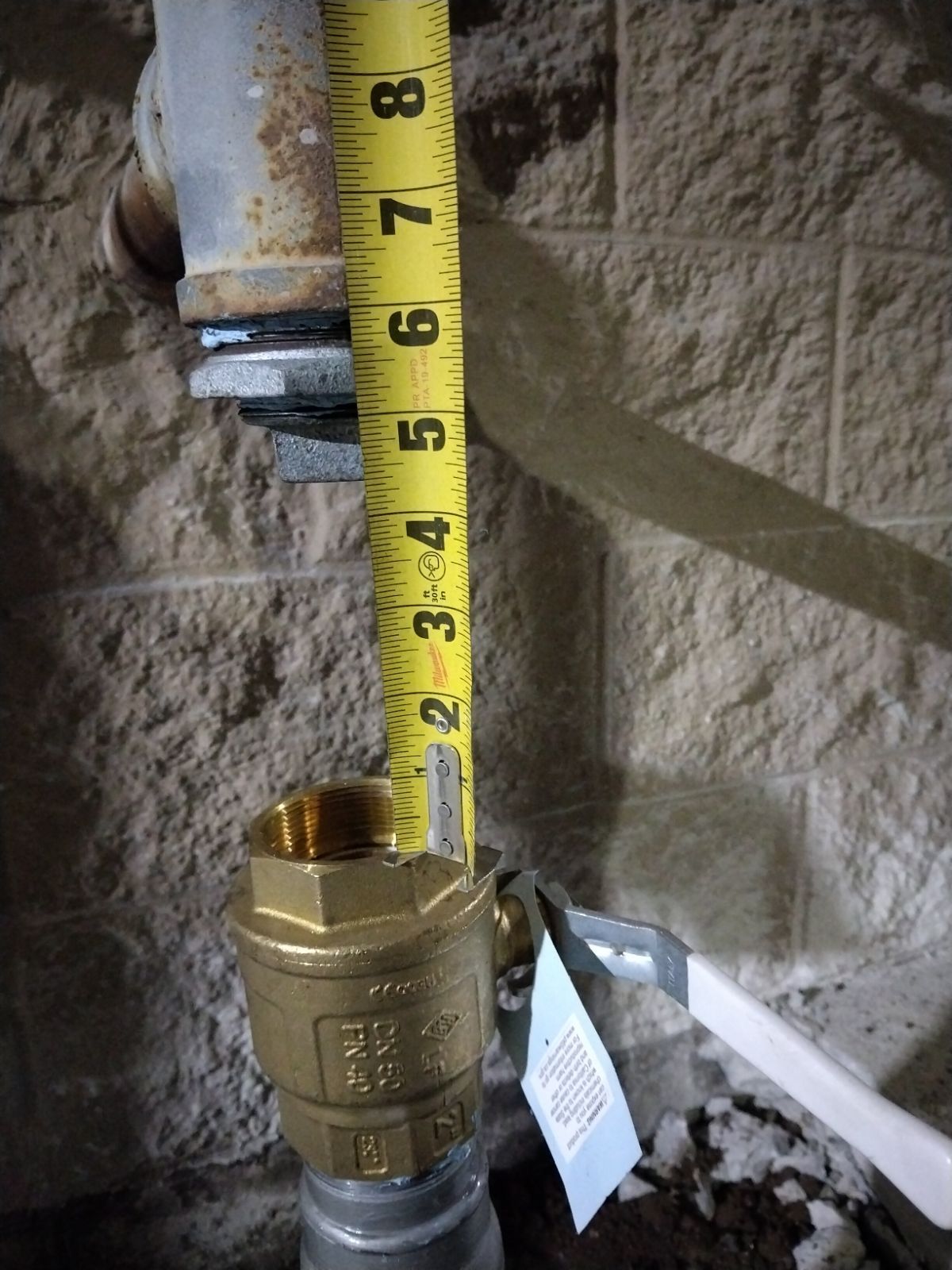 Yellow measuring tape beside a brass pipe fitting against a rough beige wall