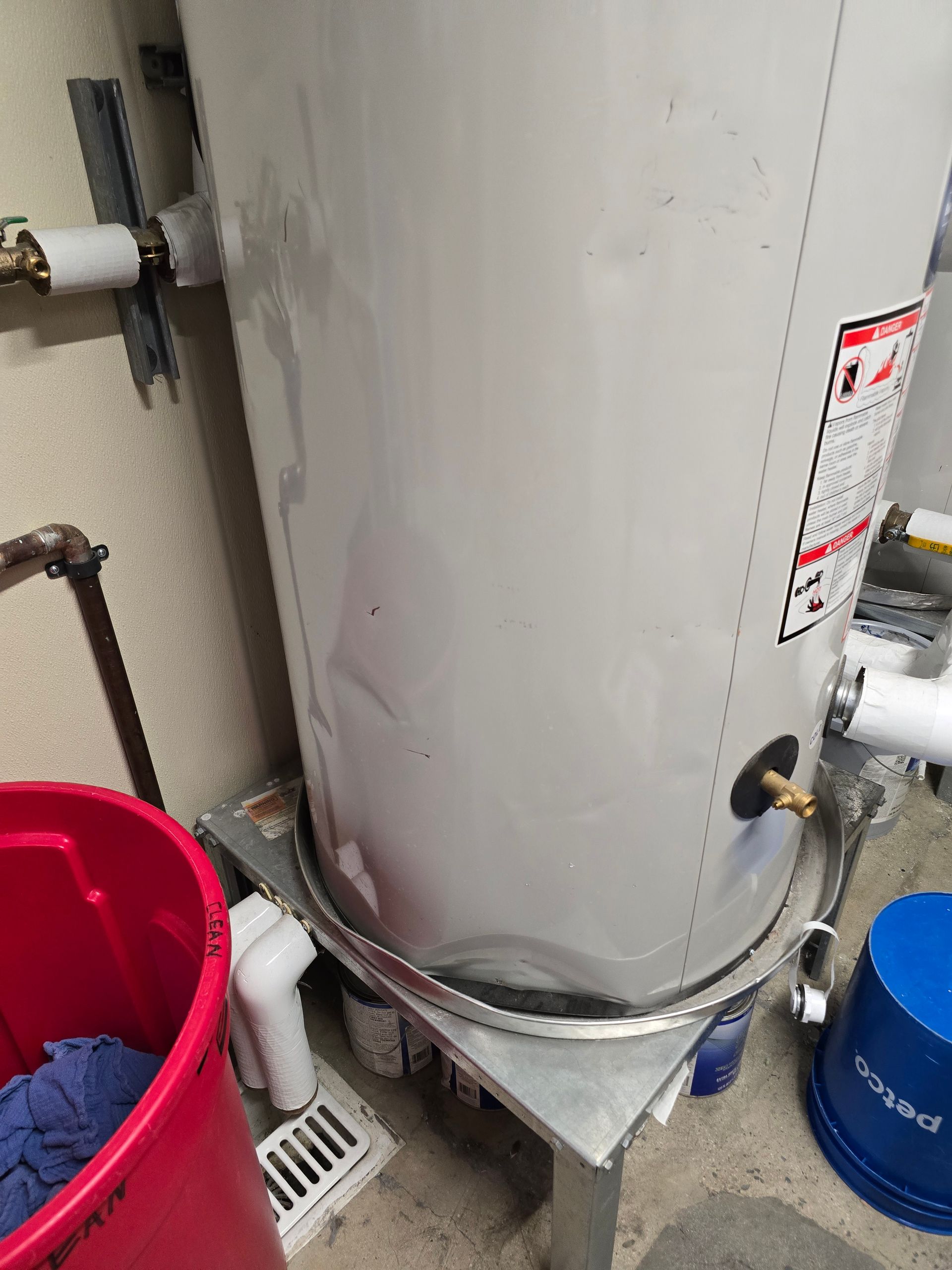White water heater with pipes and labels beside a red bucket and blue bucket in a utility area