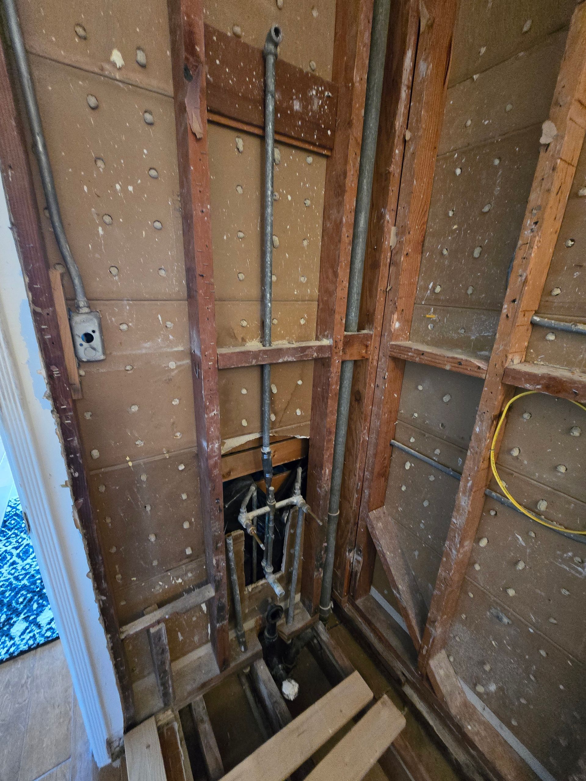 Unfinished bathroom wall framing with exposed pipes, wiring, and open floor joists during renovation