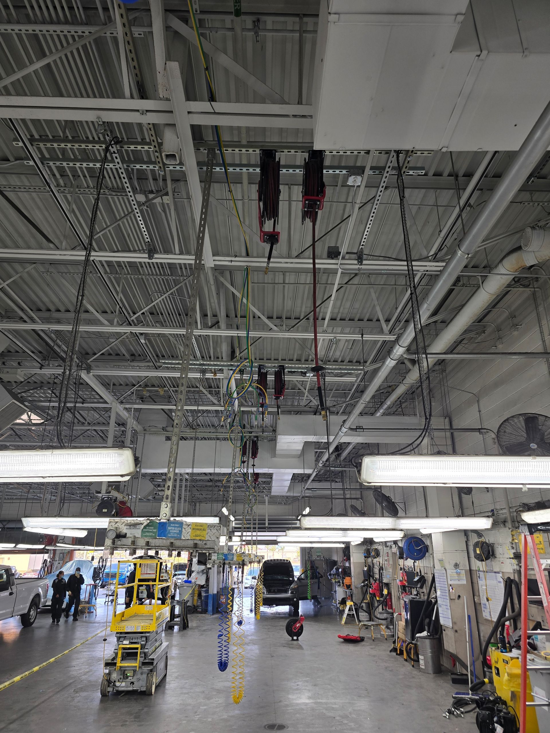 Industrial warehouse floor with hanging machinery, workstations, and carts beneath a high metal ceiling.