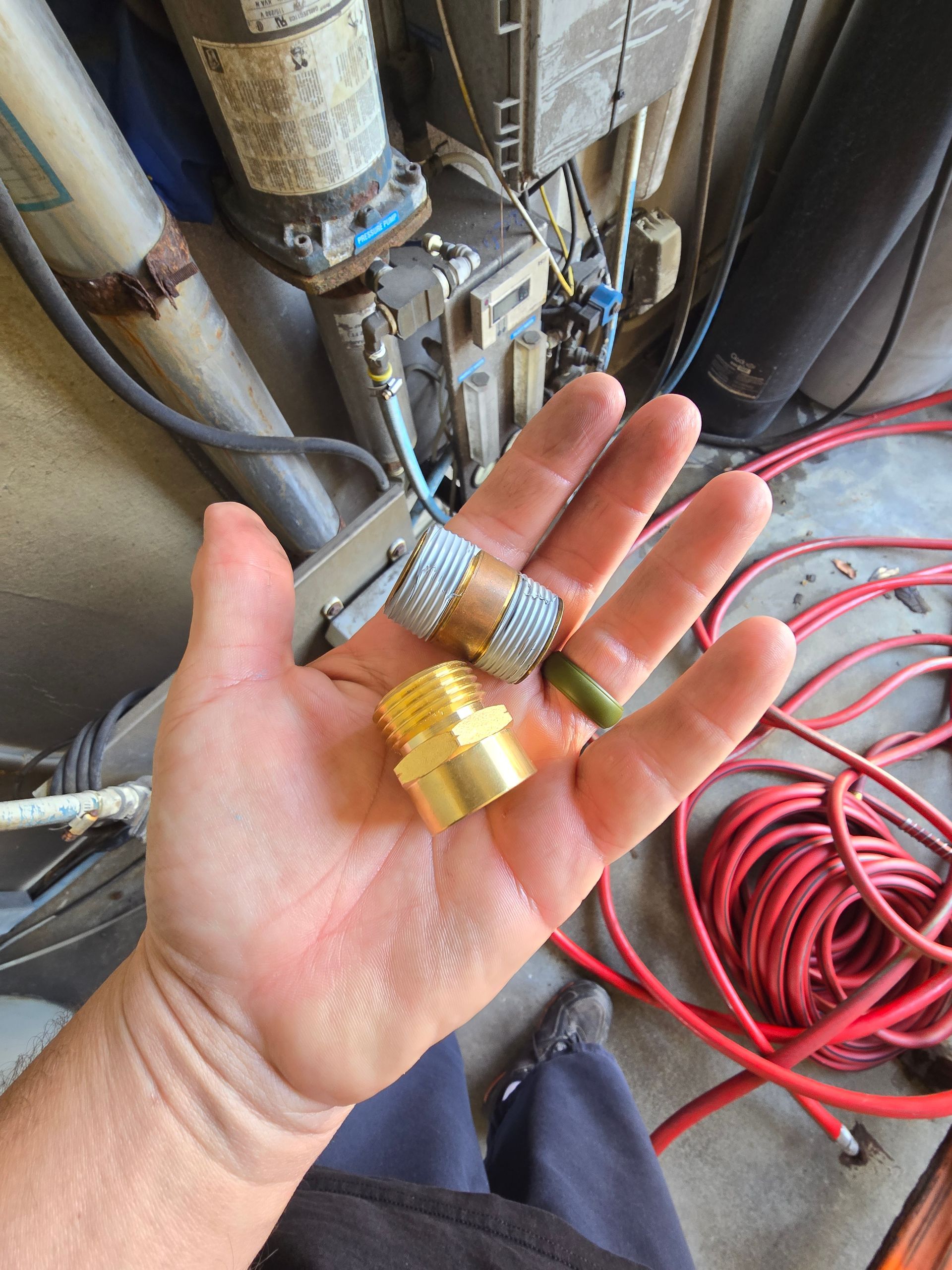 Hand holding two brass hose fittings beside pipes and coiled red hose in a workshop area