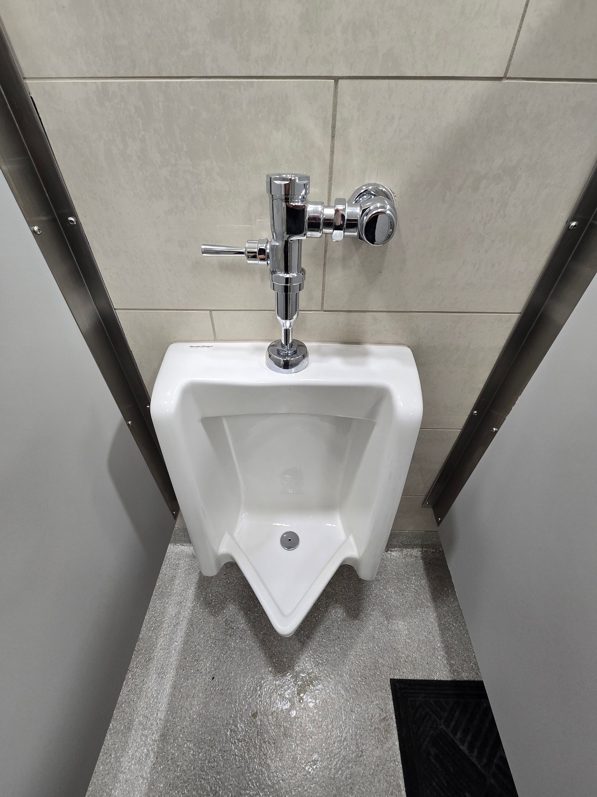Wall-mounted urinal in a tiled restroom stall with a chrome flush valve and gray speckled floor.