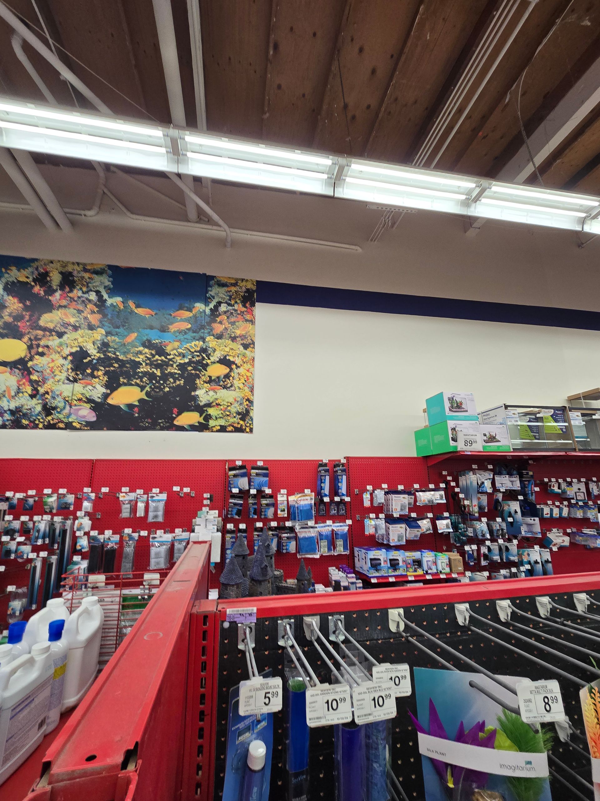 A store aisle with red display racks, hanging merchandise, and fluorescent ceiling lights beneath a floral wall art panel