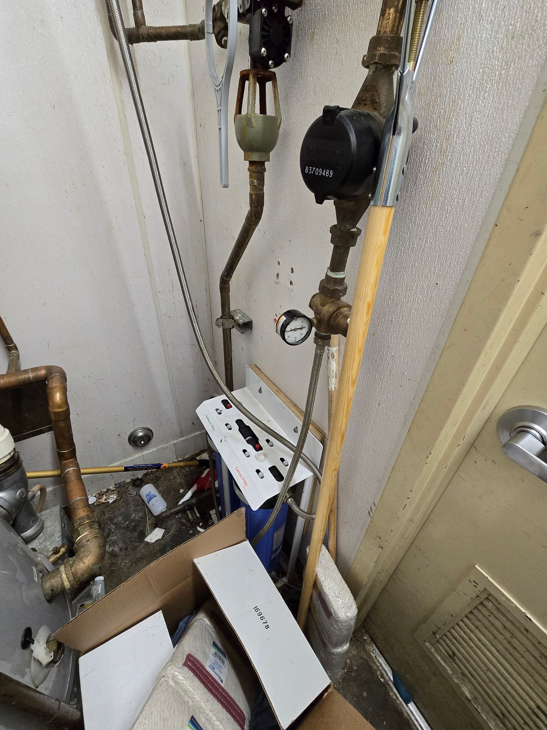 Cluttered utility closet with exposed pipes, valves, and a door on the right.