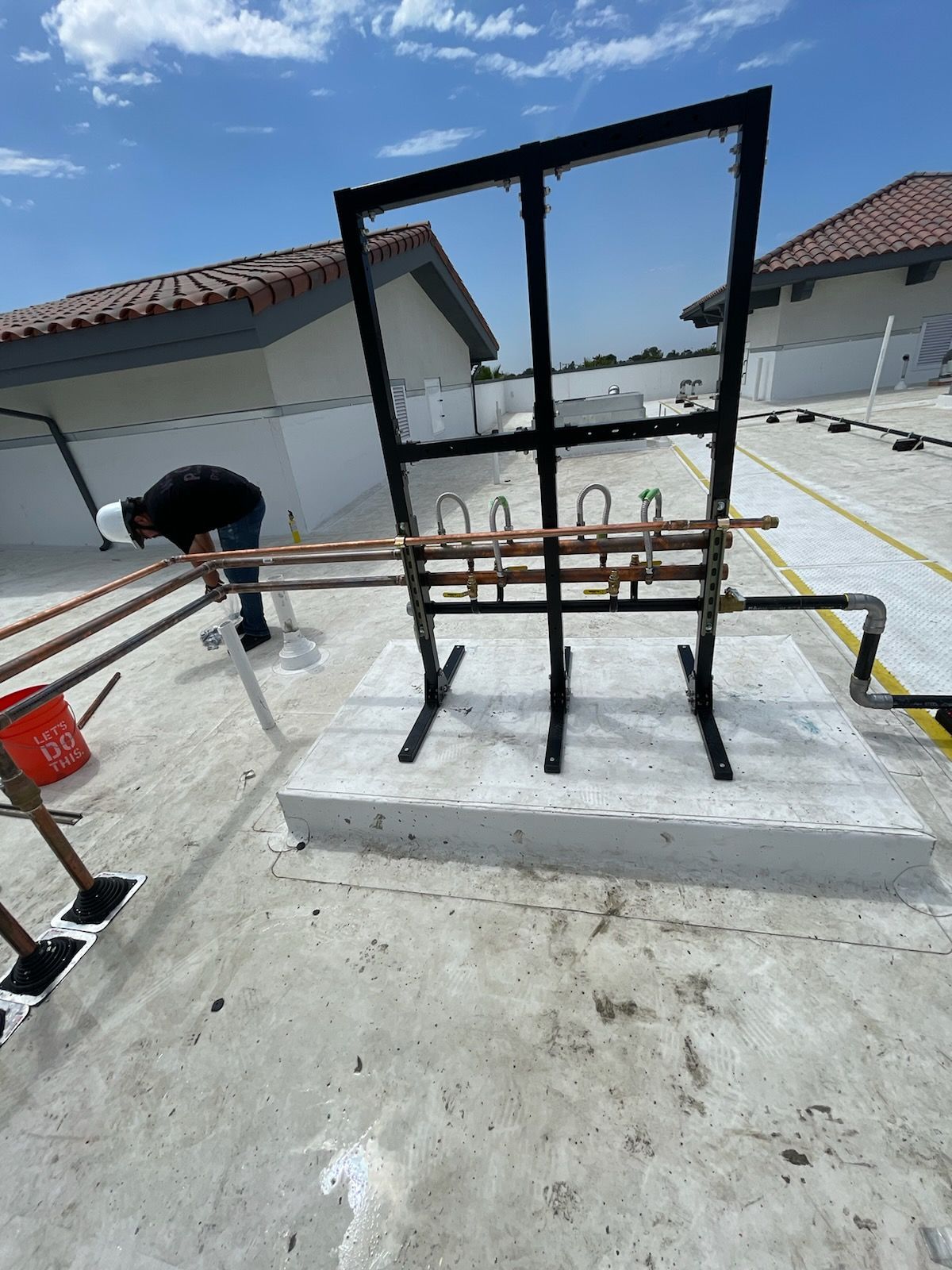 Black metal frame on a rooftop concrete pad, with pipes and a person bending over on the left.