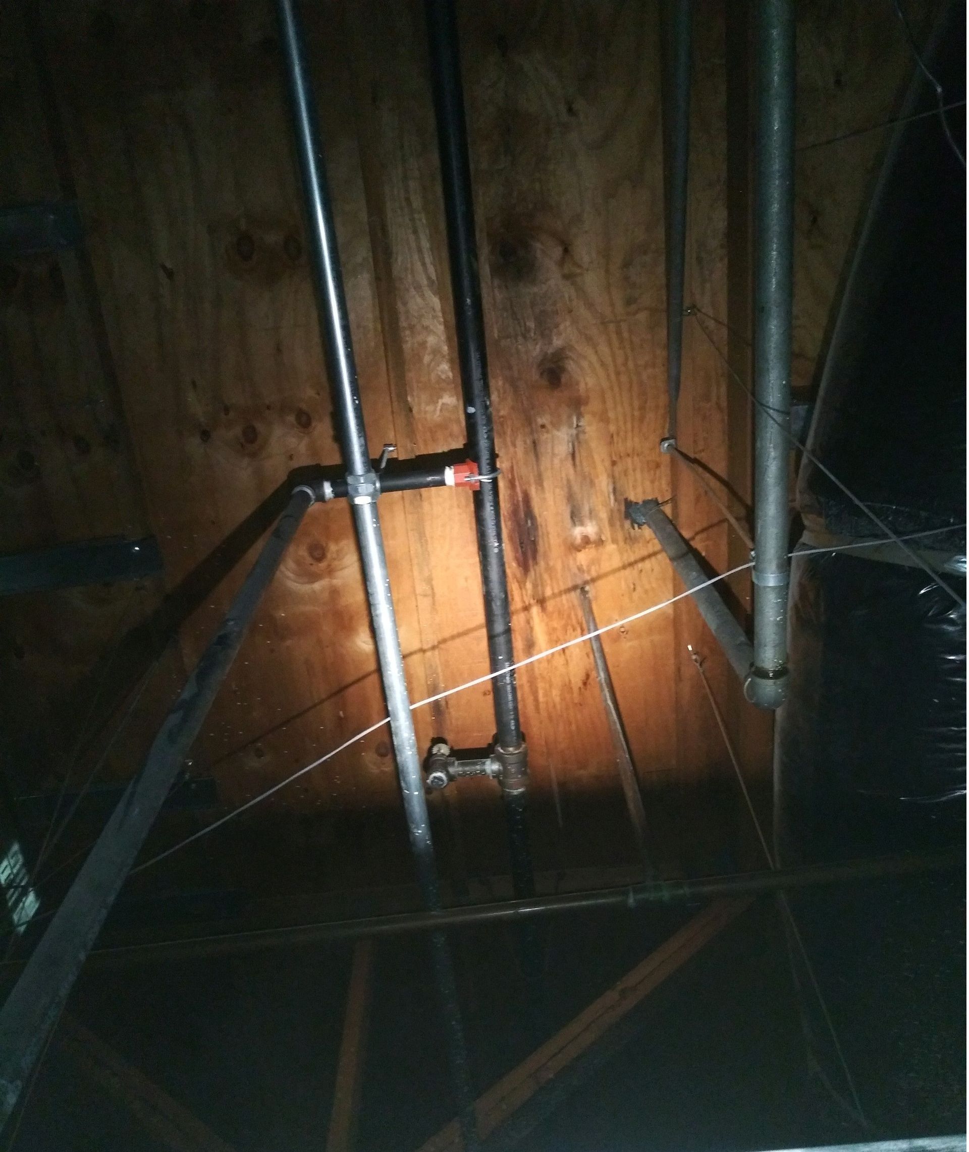 Exposed wooden wall with metal pipes and wiring in a dim attic or crawlspace