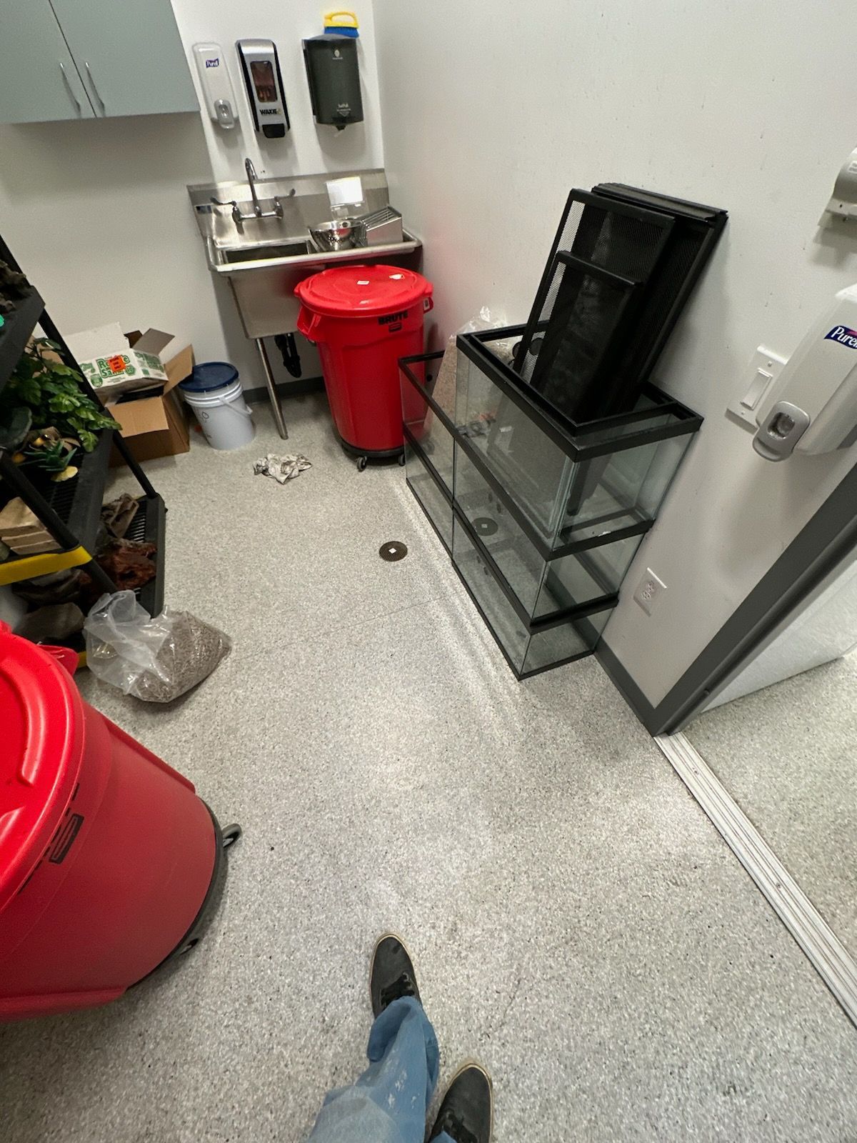 Small utility room with red bins, sink, cleaning supplies, and stacked black trays on a cart.