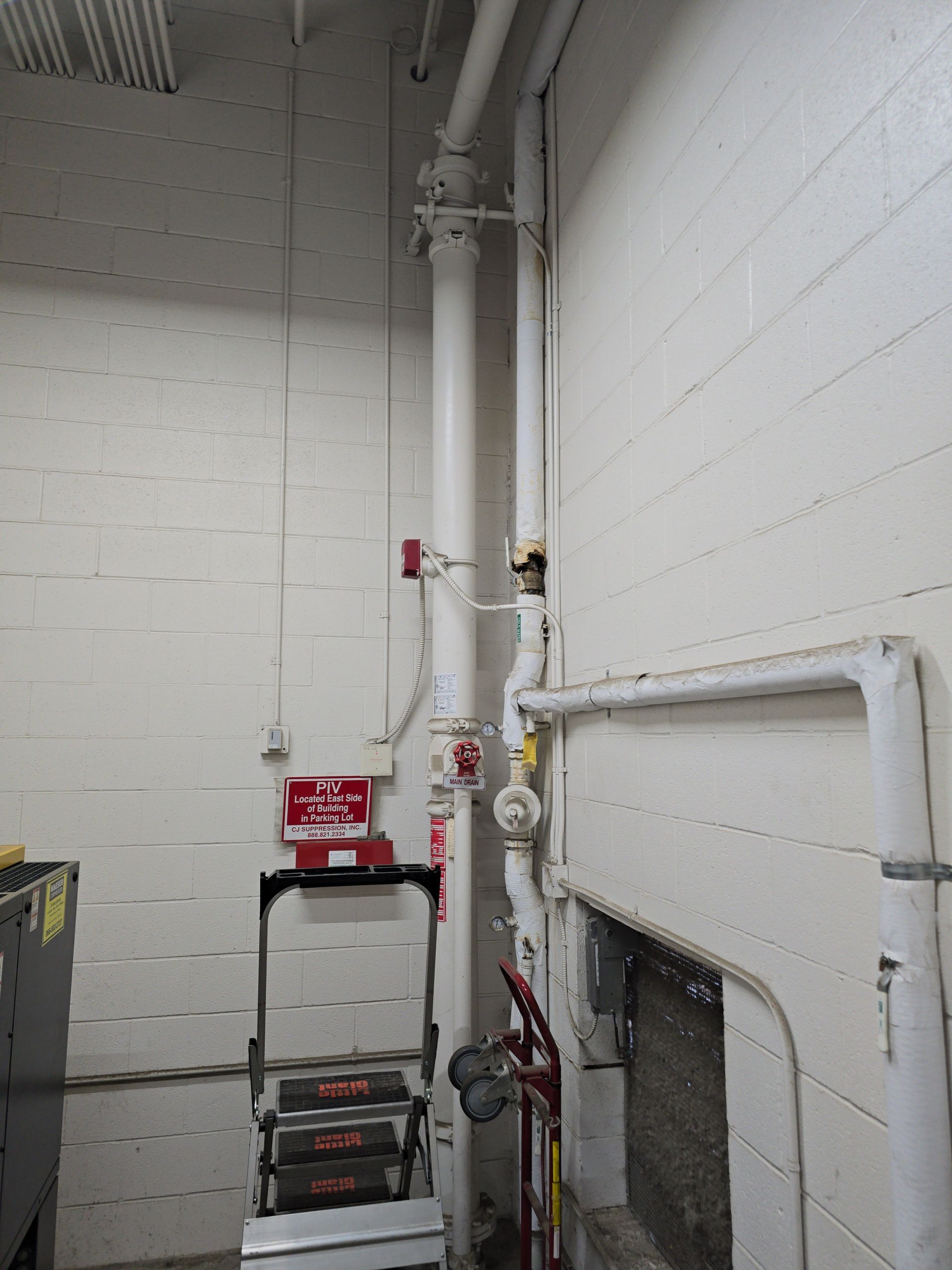 Industrial utility room with white cinderblock walls, pipes, and a red ladder cart near a wall-mounted fixture
