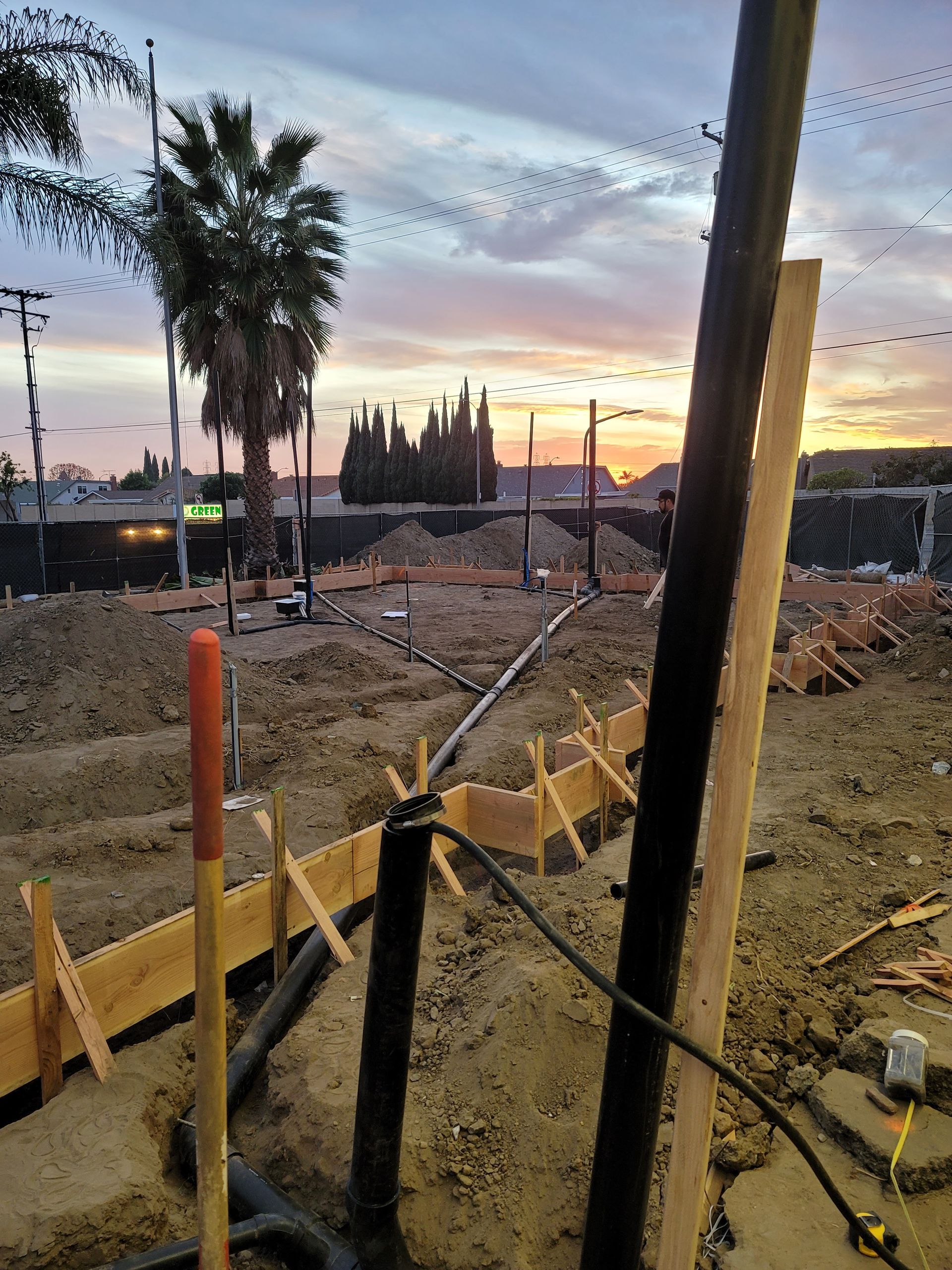Construction site at sunset with dirt excavation, wooden forms, and utility poles under a pink sky