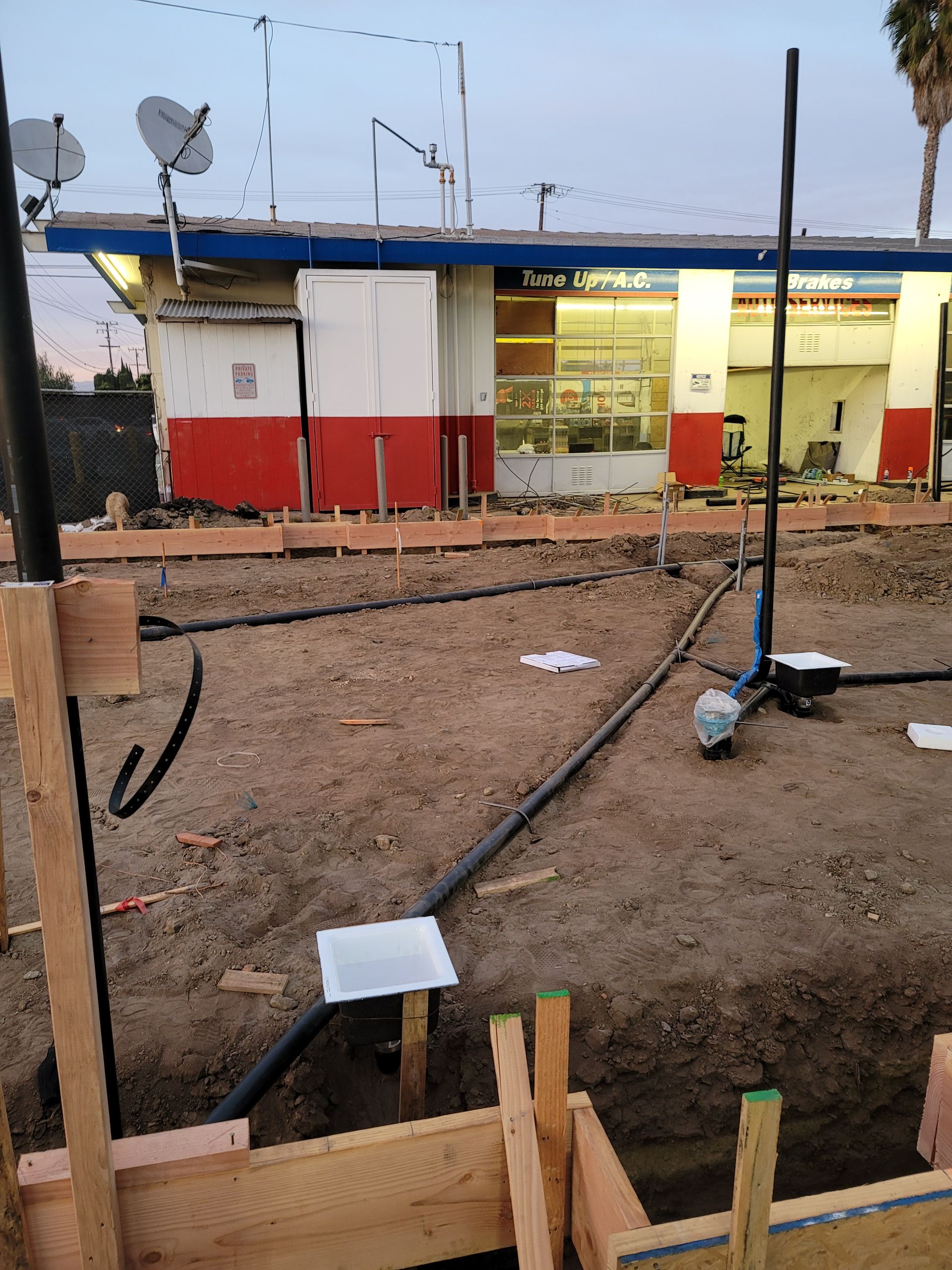 Trench in a construction site with hoses and wooden supports beside a small red-and-white building at dusk