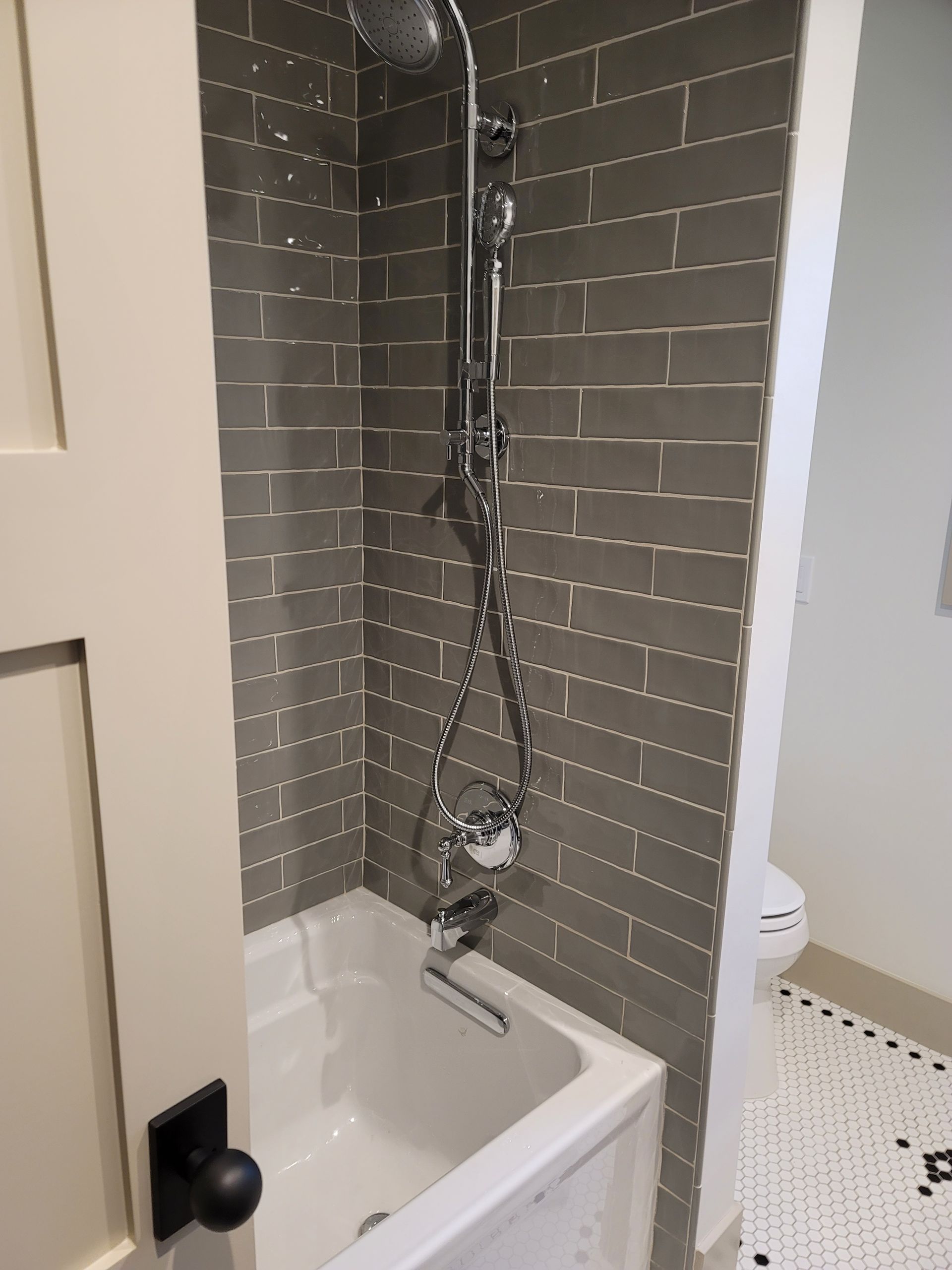 Small bathroom shower with gray tile walls, white tub, and exposed shower plumbing.