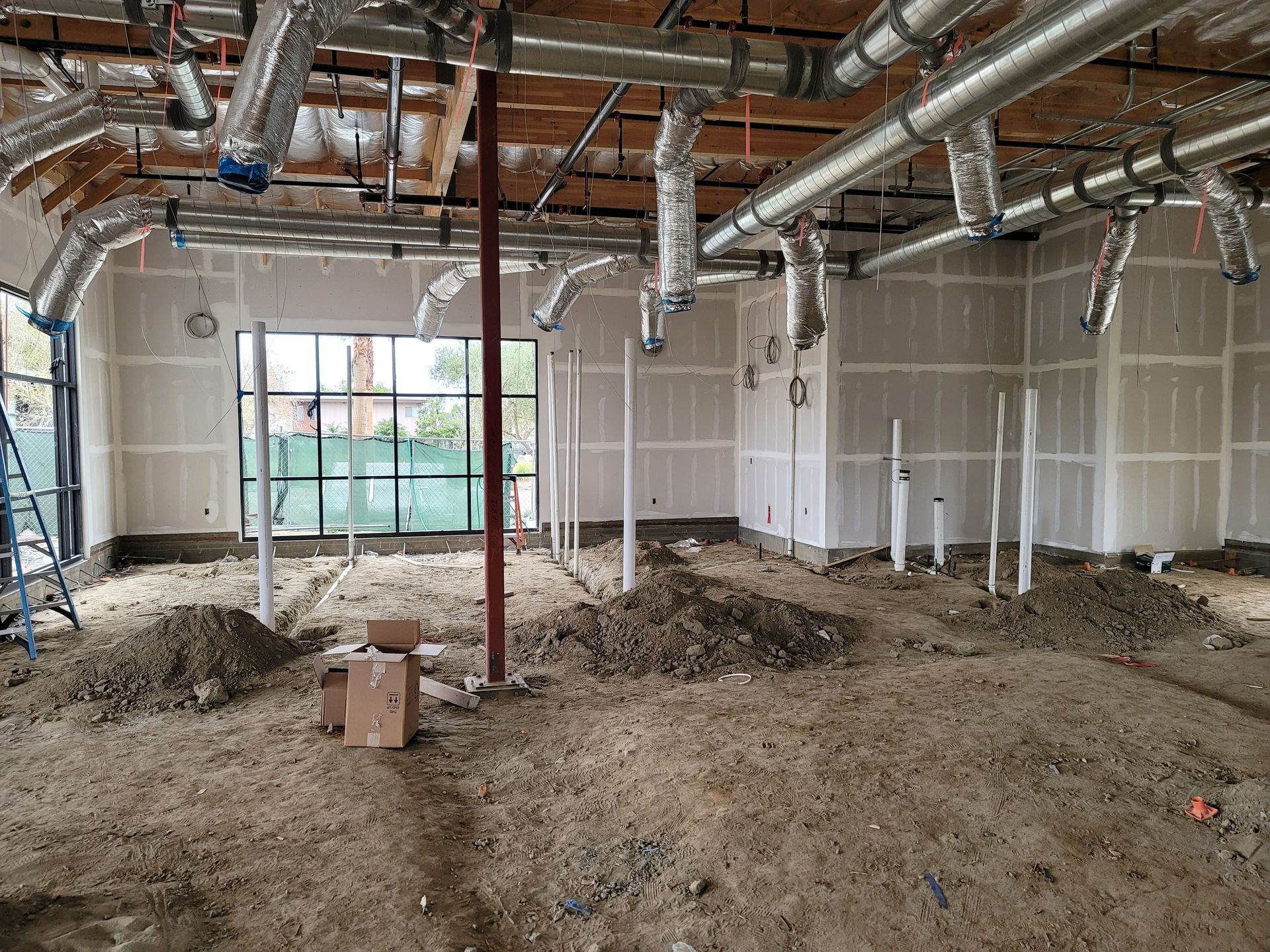 Unfinished office interior with exposed ductwork, drywall, dirt floor, and large windows.