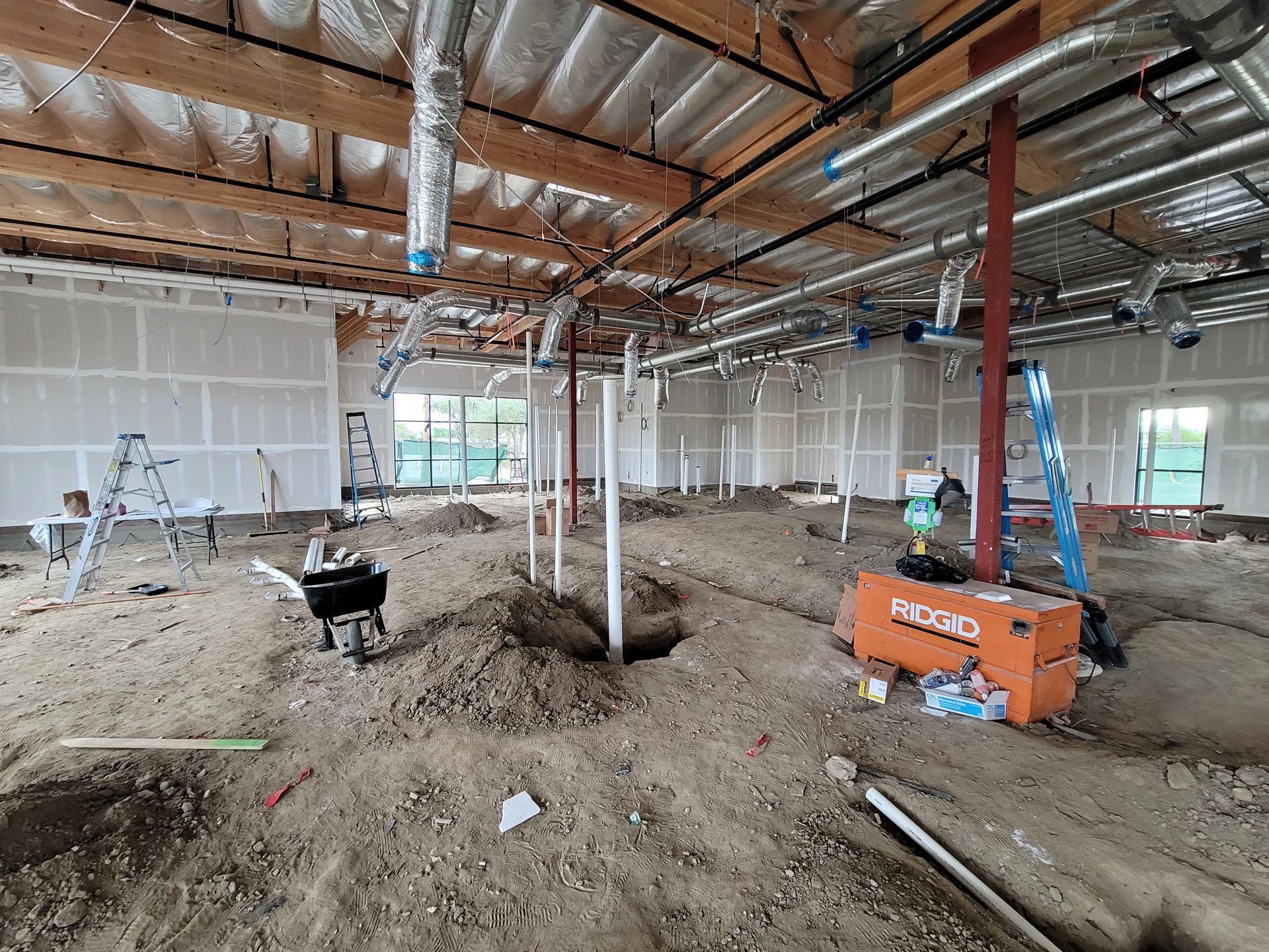 Construction site interior with exposed beams, drywall, ladders, and equipment on a dirt floor