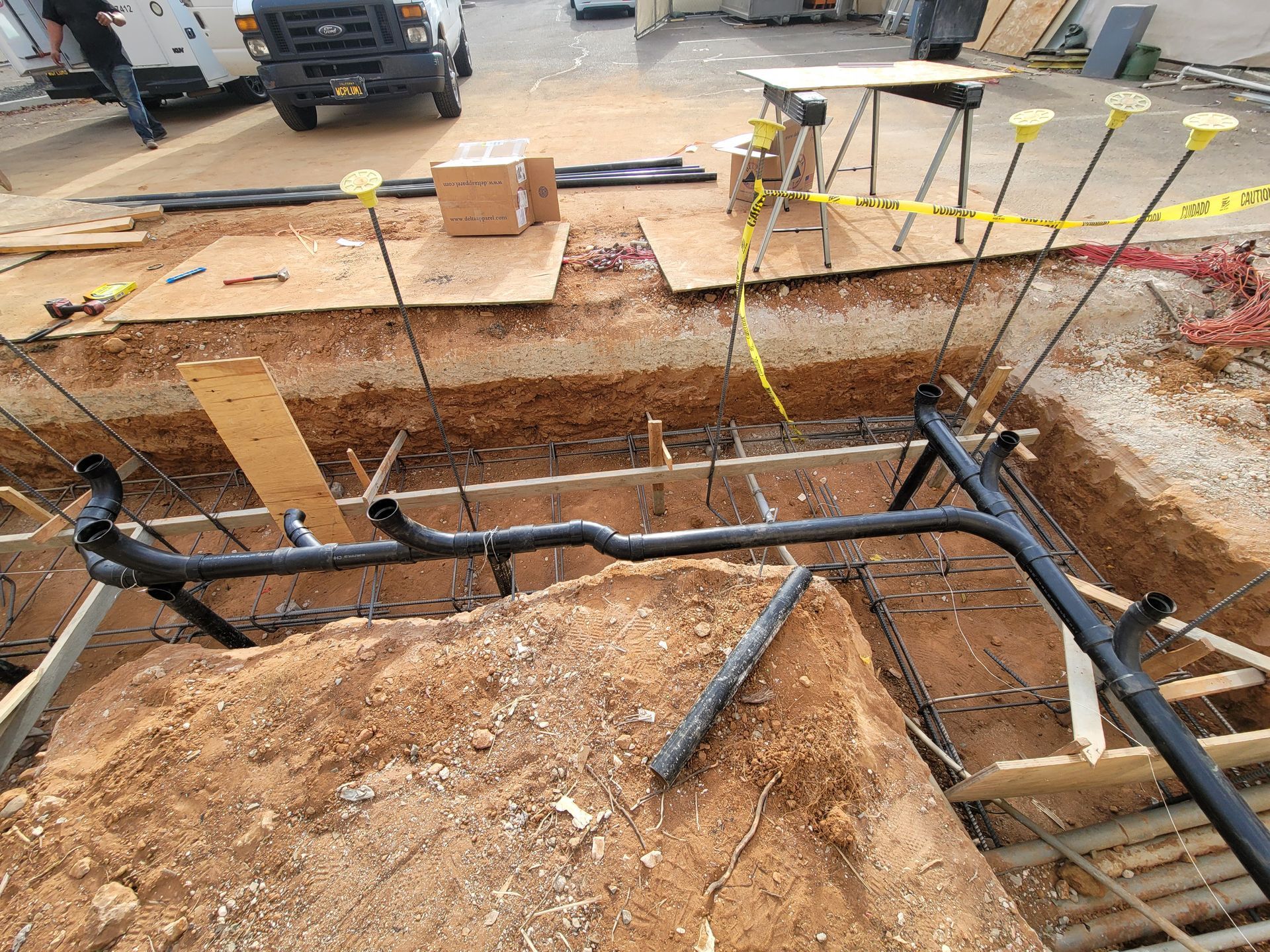 Excavation site with exposed black pipes, red dirt trench, and yellow caution tape near a street.