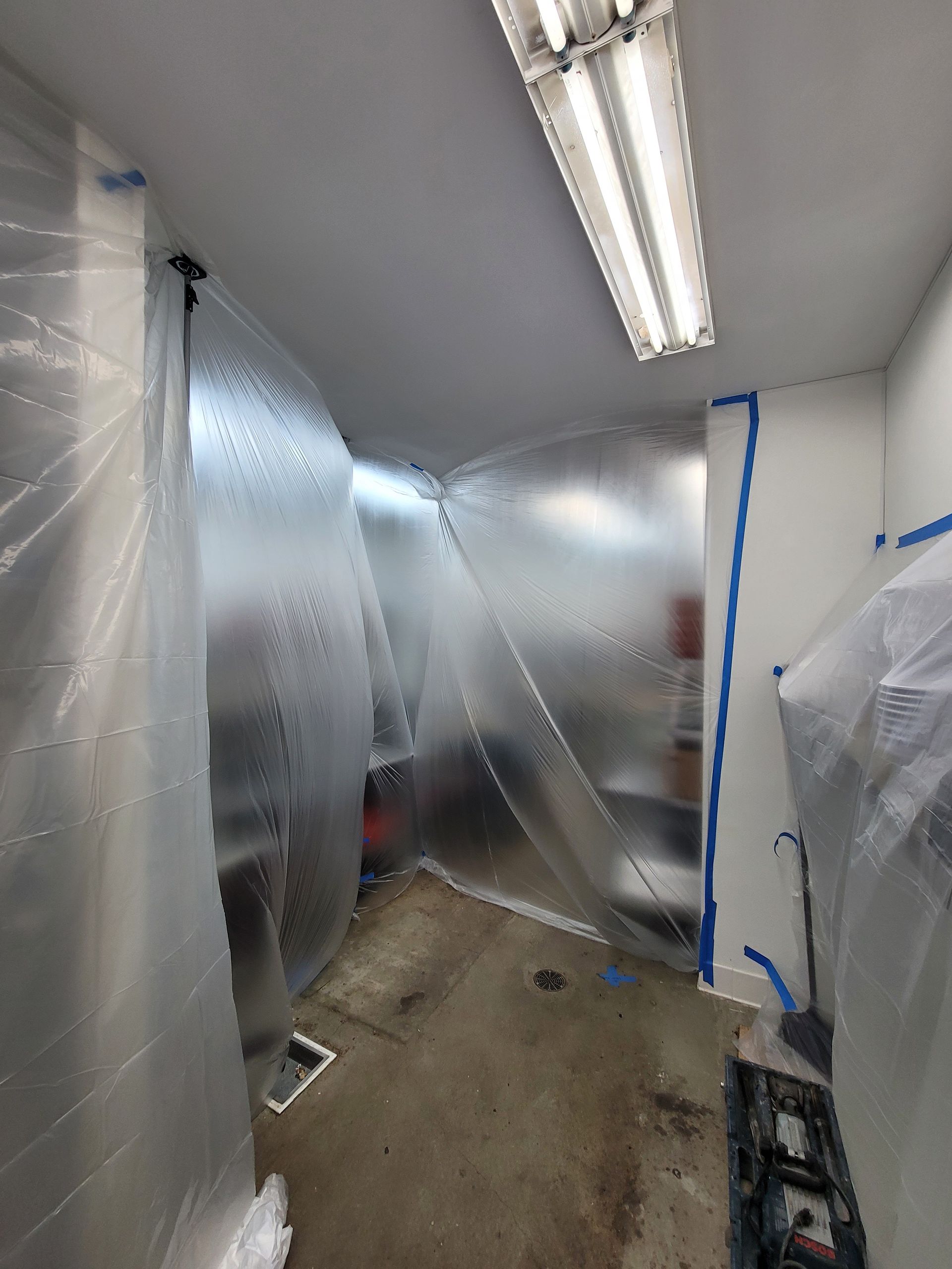 Plastic-covered hallway under fluorescent lights with a concrete floor and blue tape on a doorway