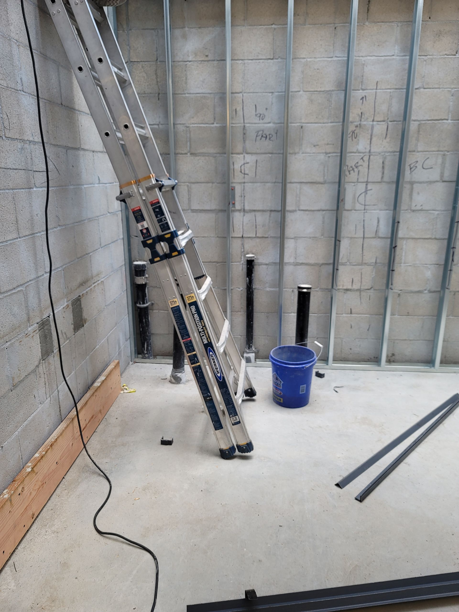 Ladder propped in a concrete basement corner beside a blue bucket and two metal rods on the floor
