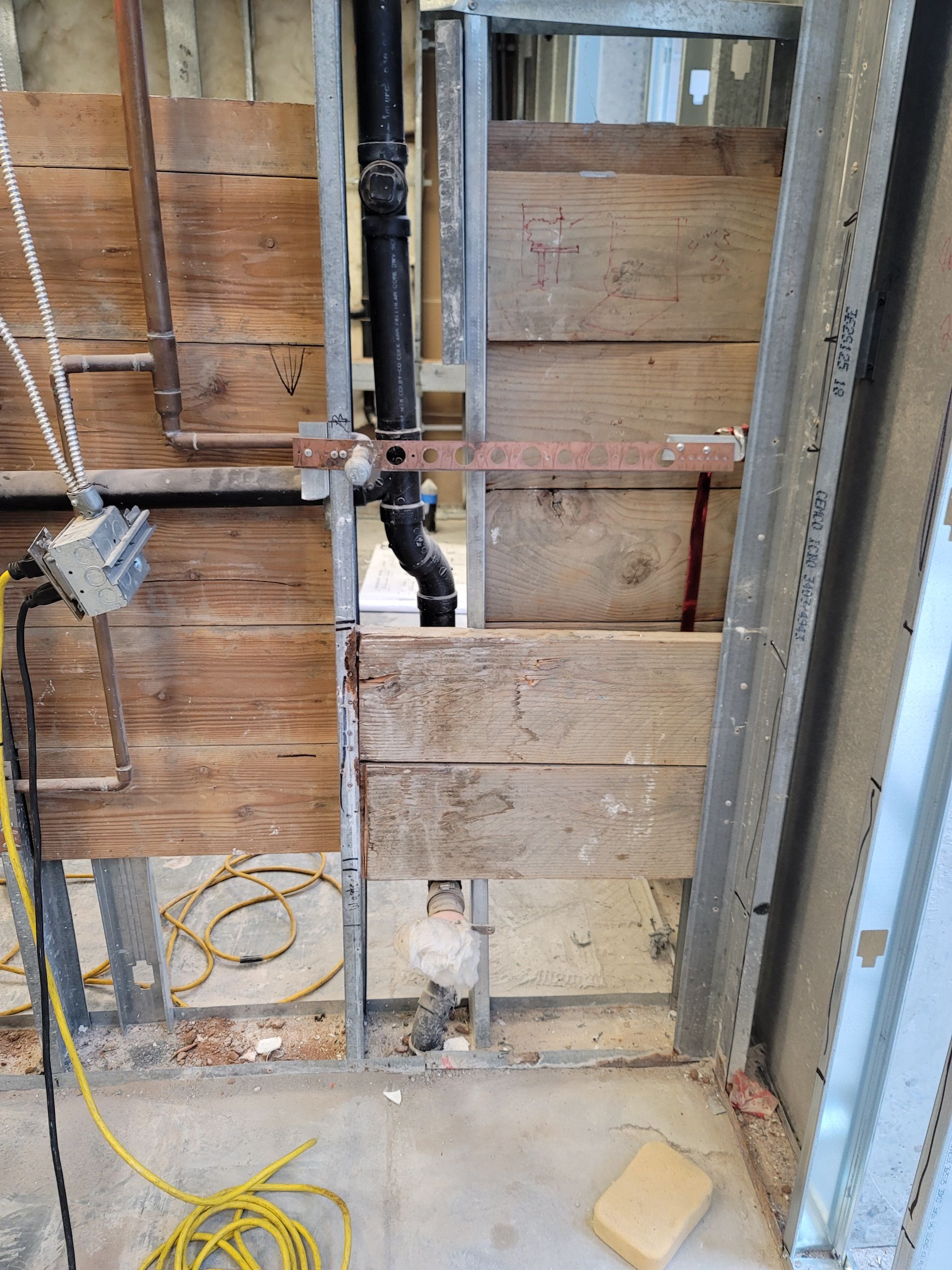 Exposed wall studs and plumbing during renovation, with unfinished plywood sheathing and yellow cords on the floor.