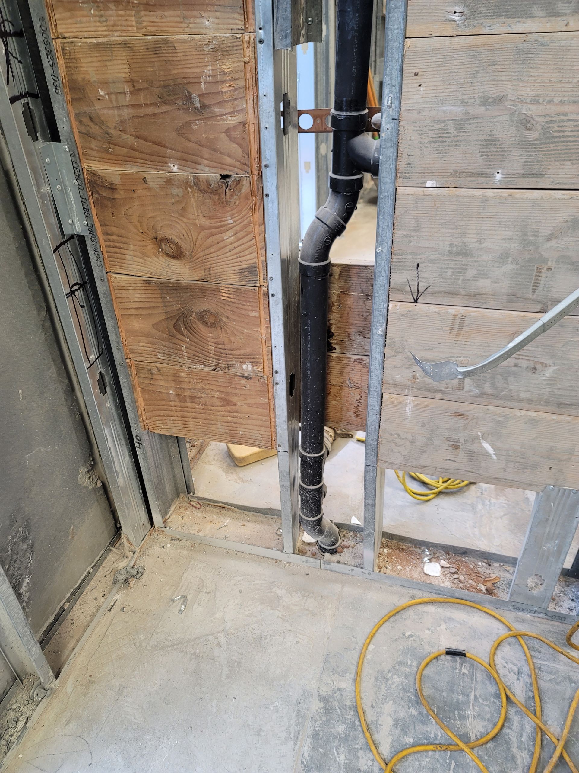 Partially opened wall with exposed plumbing pipes and wiring in a construction area with dusty floor and coiled yellow cord