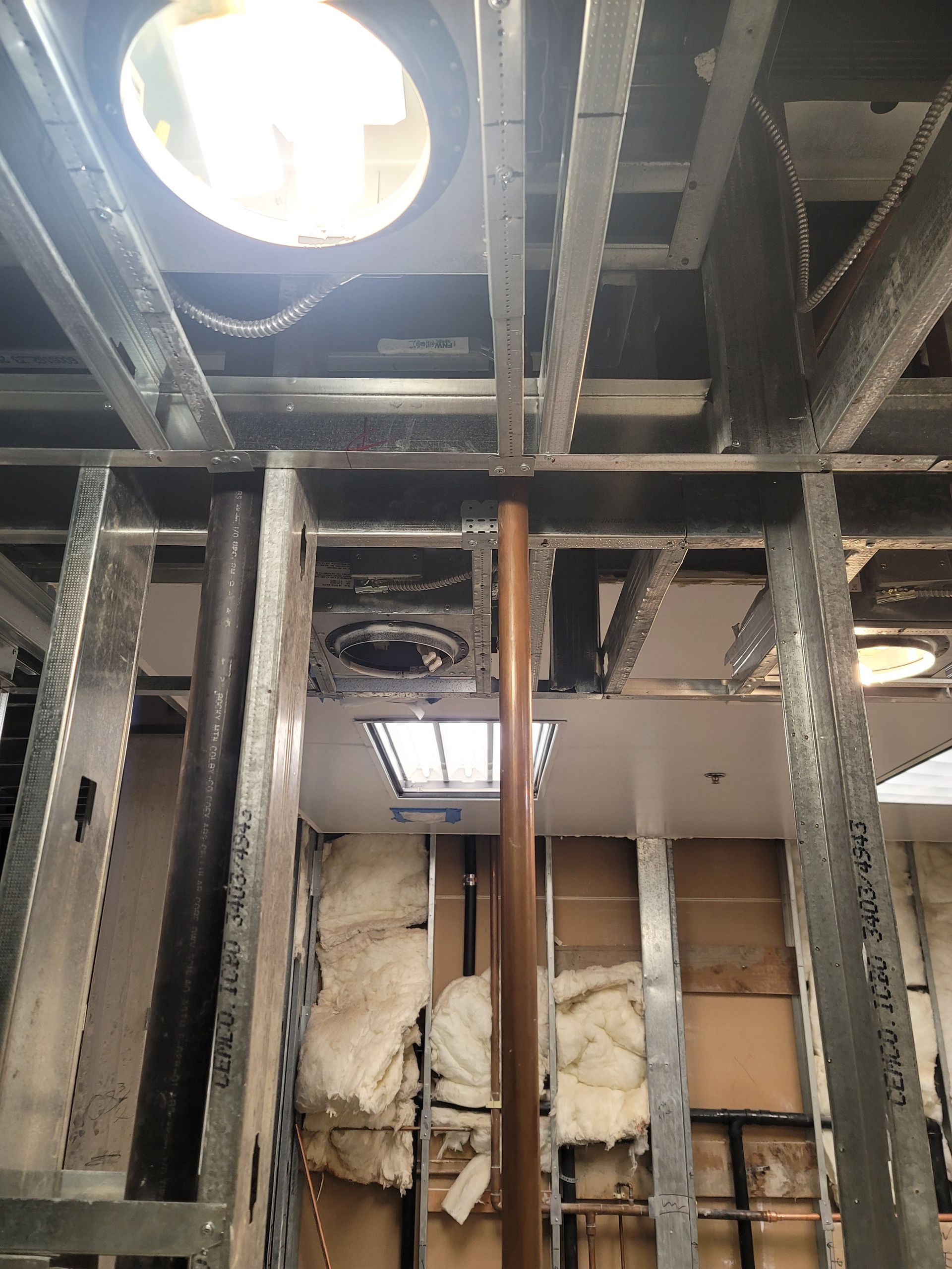 Exposed ceiling framing and insulation in a room under renovation, with a round skylight and fluorescent light panel.