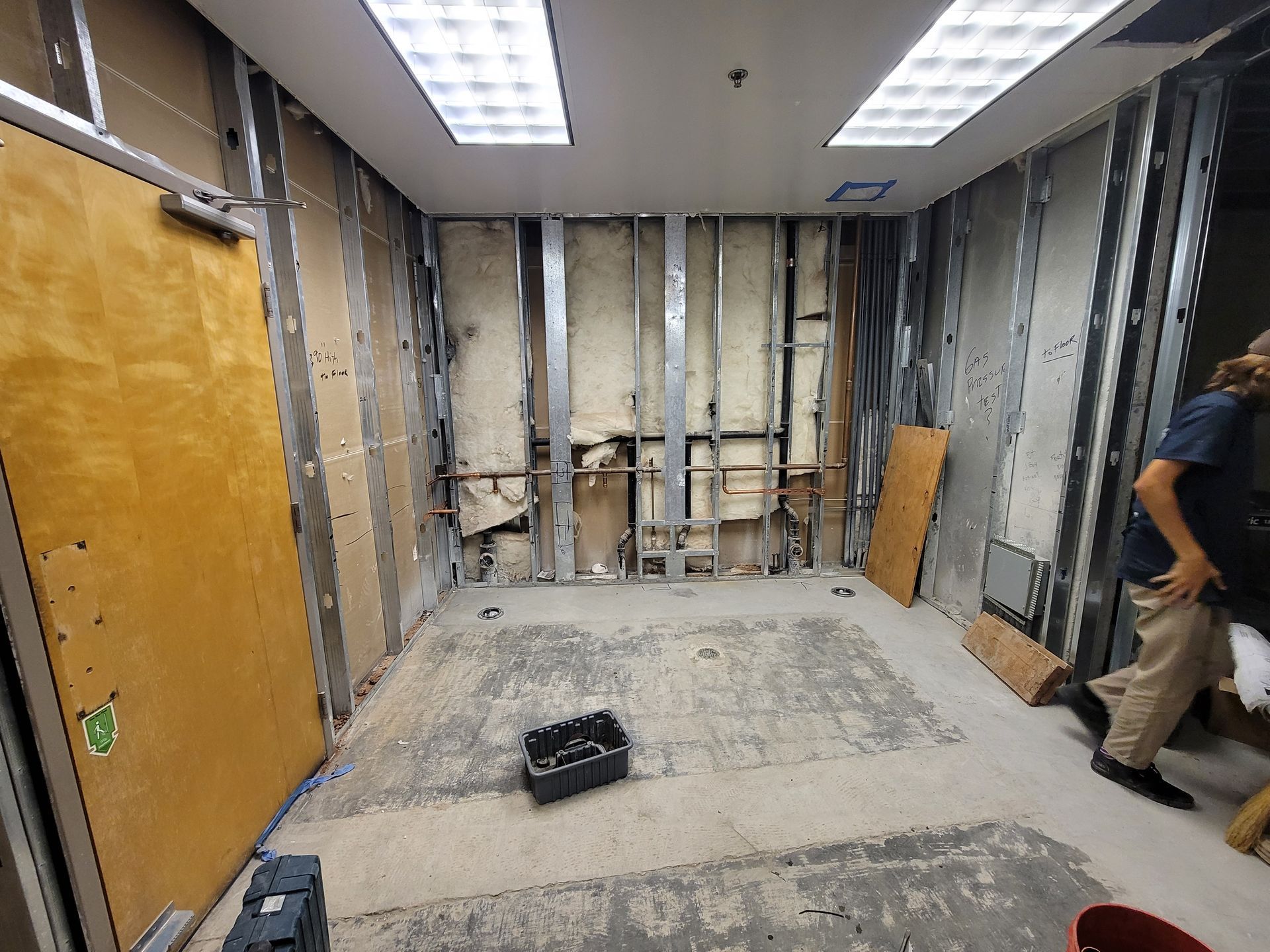 Bare office room under renovation, with exposed studs, fluorescent lights, and construction tools on the floor.