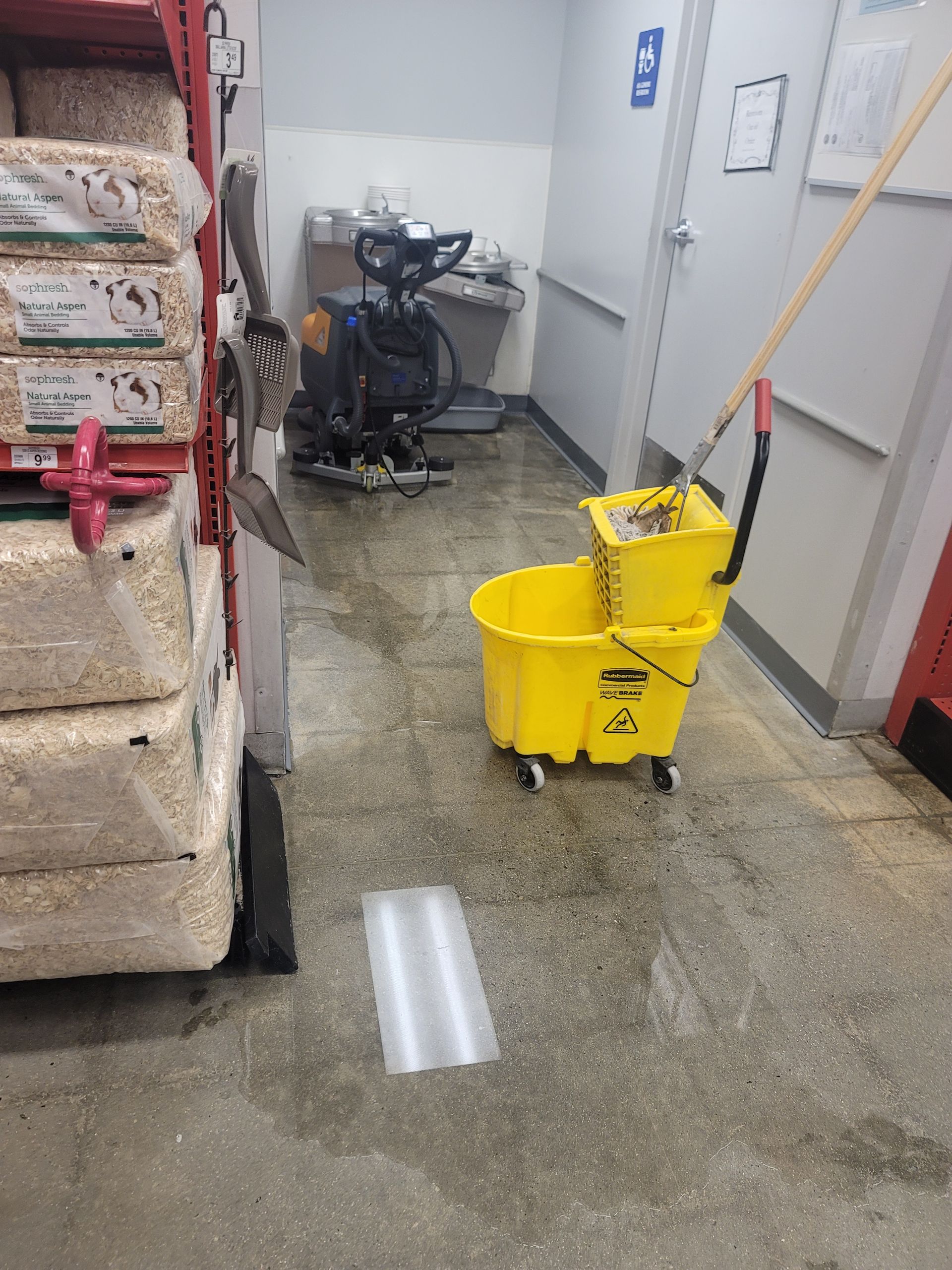 Mop bucket and wringer on a wet hallway floor beside stacked supplies and a utility cart