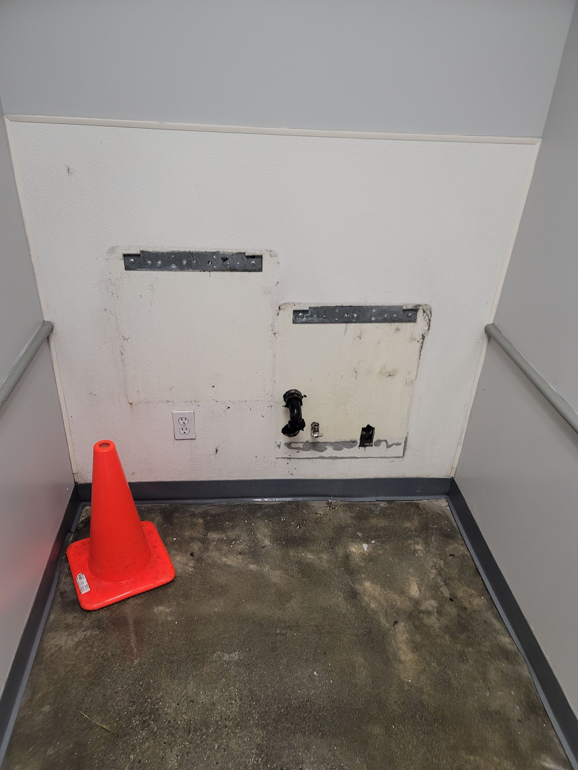 Small utility alcove with a white wall, two access panels, a cone, and a worn floor.