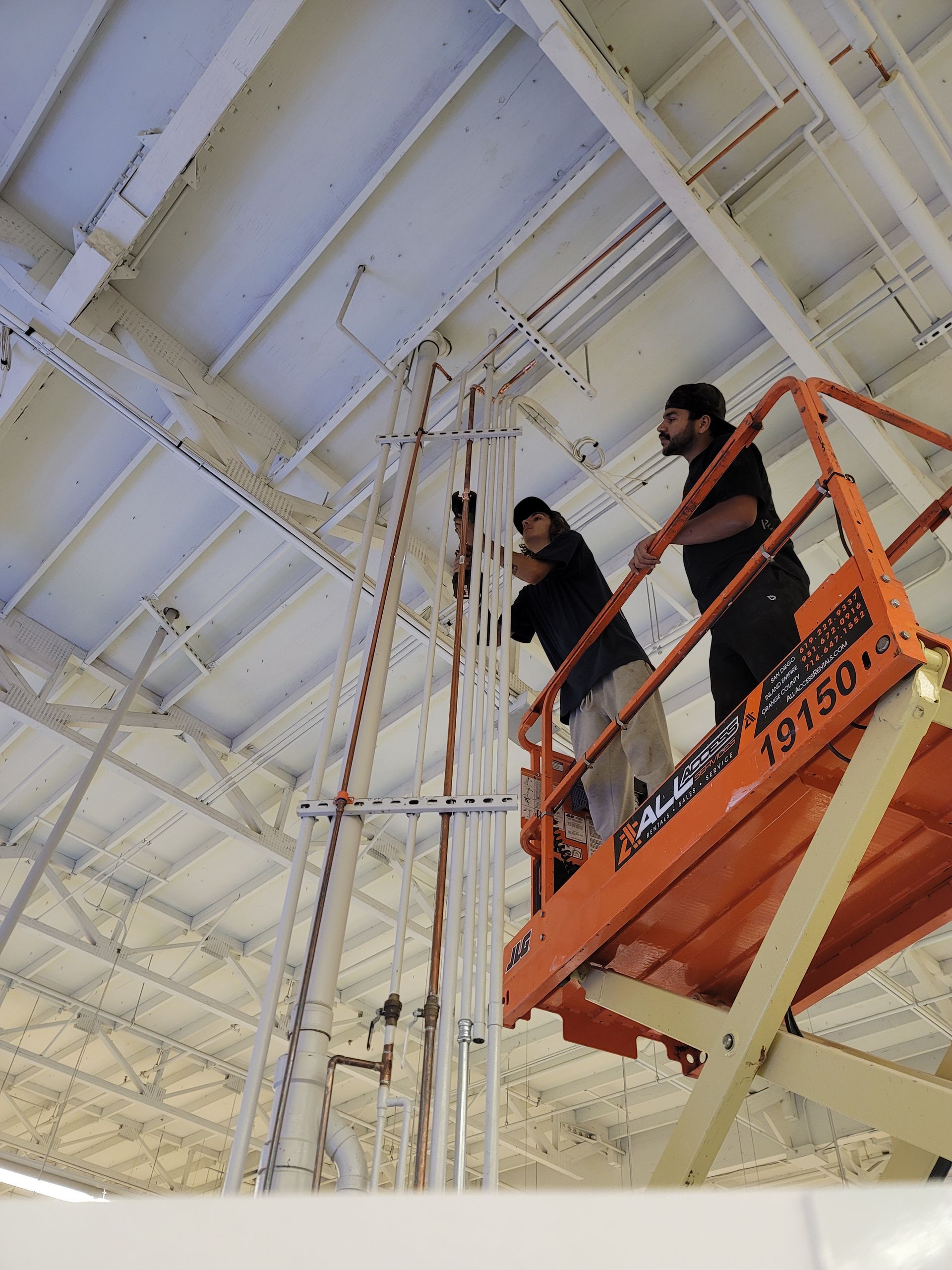 Two workers on an orange lift inspect the ceiling inside a bright industrial building