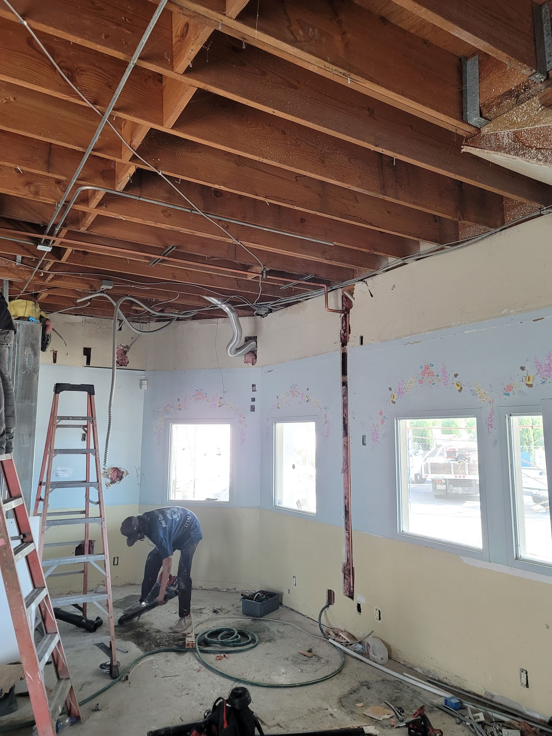 Renovation work in a room with exposed ceiling beams, ladders, and debris-covered floor.