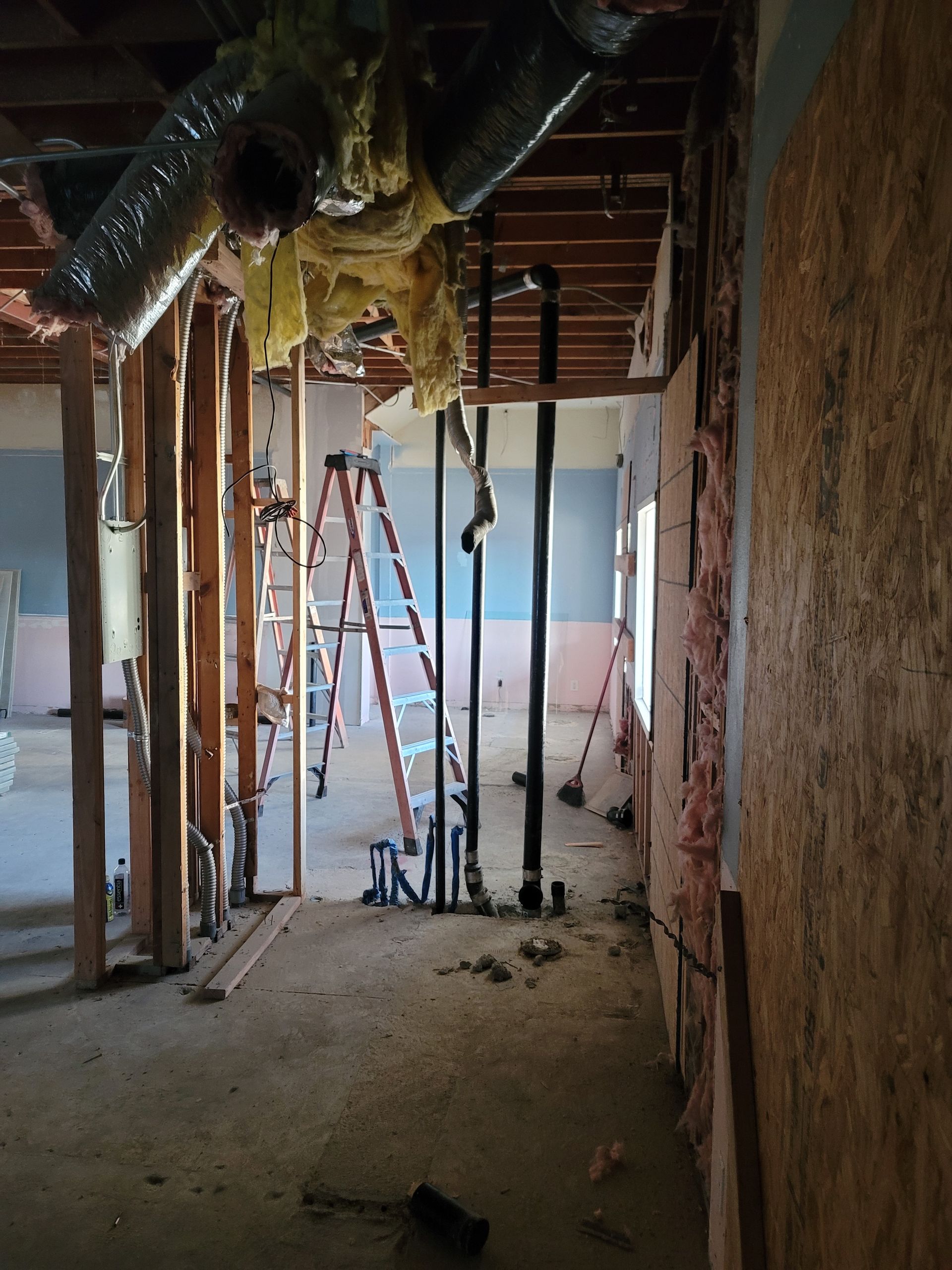 Partially demolished interior with exposed studs, pipes, debris, and a ladder in the background