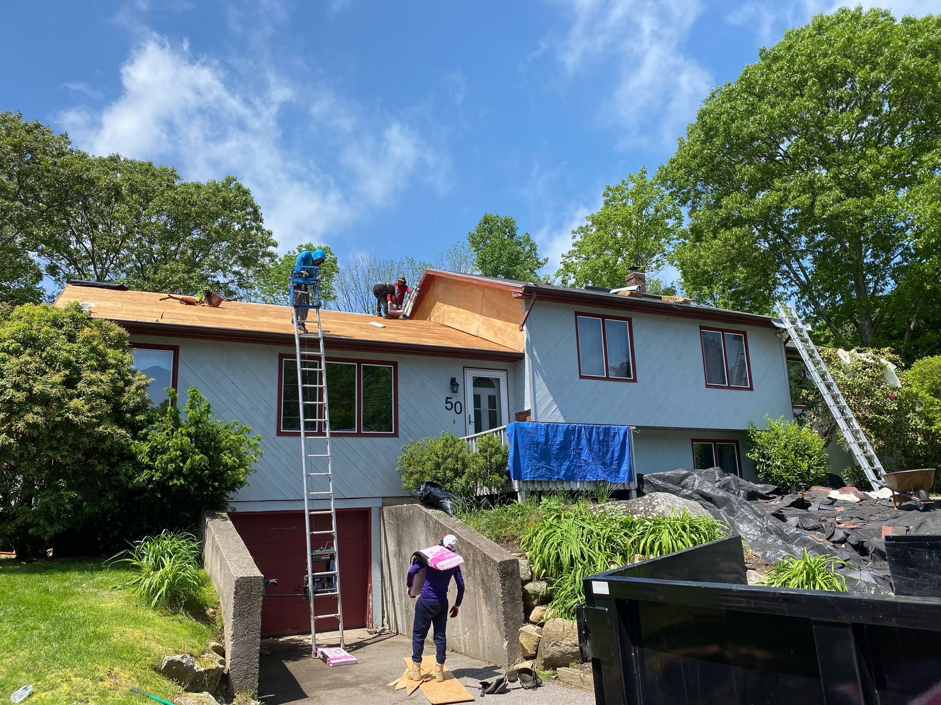 A house under renovation with workers on the roof and ladders, with a dumpster and supplies in the yard.