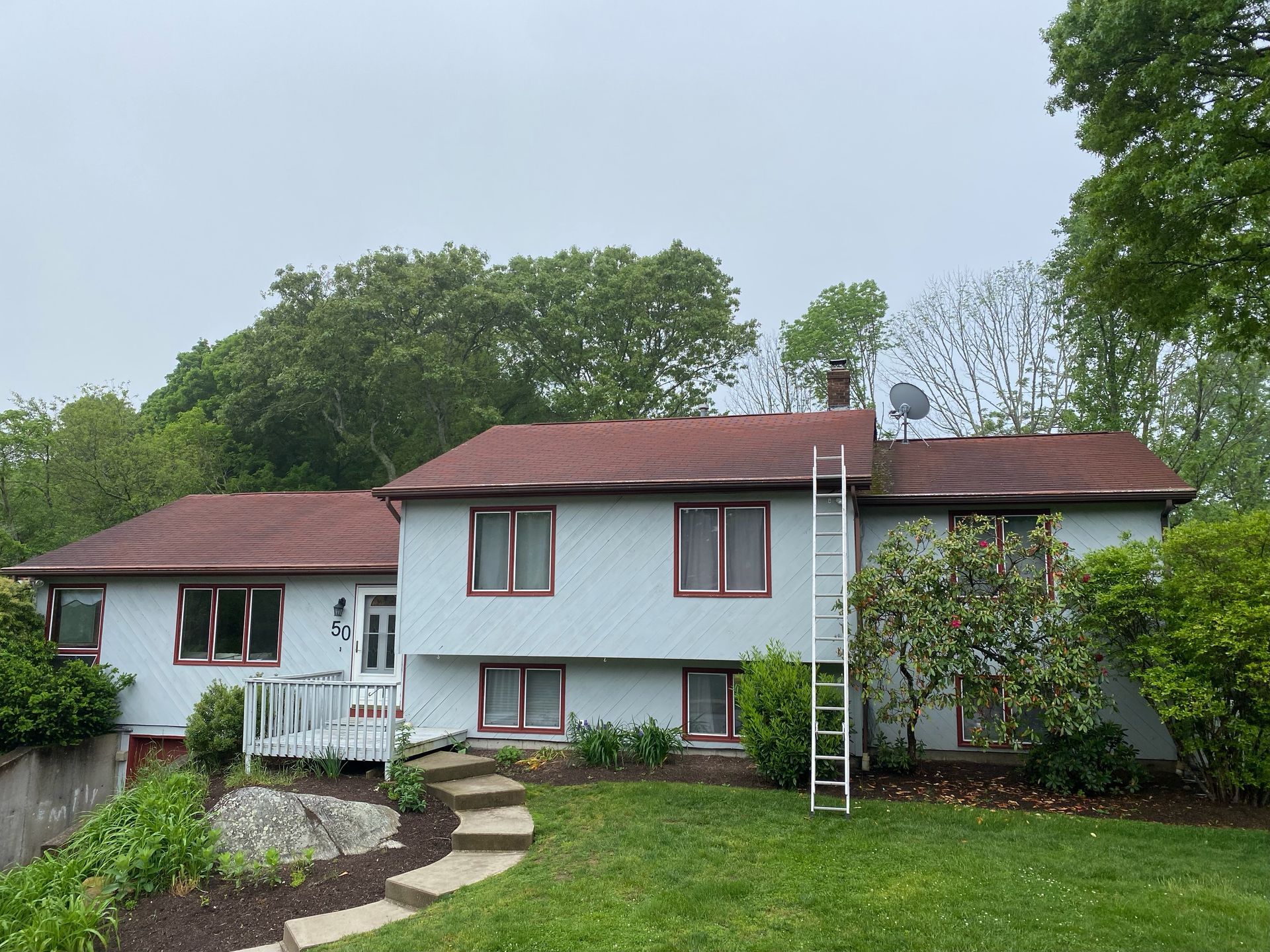 A light blue, two-story house with a reddish-brown roof, surrounded by trees and a stone-stepped path leading to a deck.