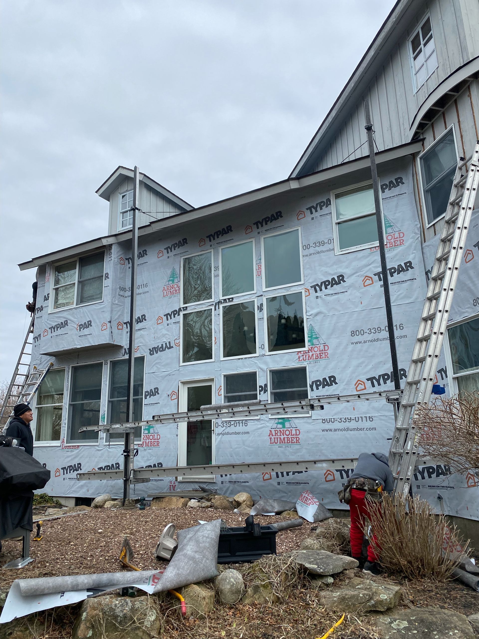 Workers on scaffolding renovate the exterior of a house covered in Typar moisture barrier wrap under a cloudy sky.
