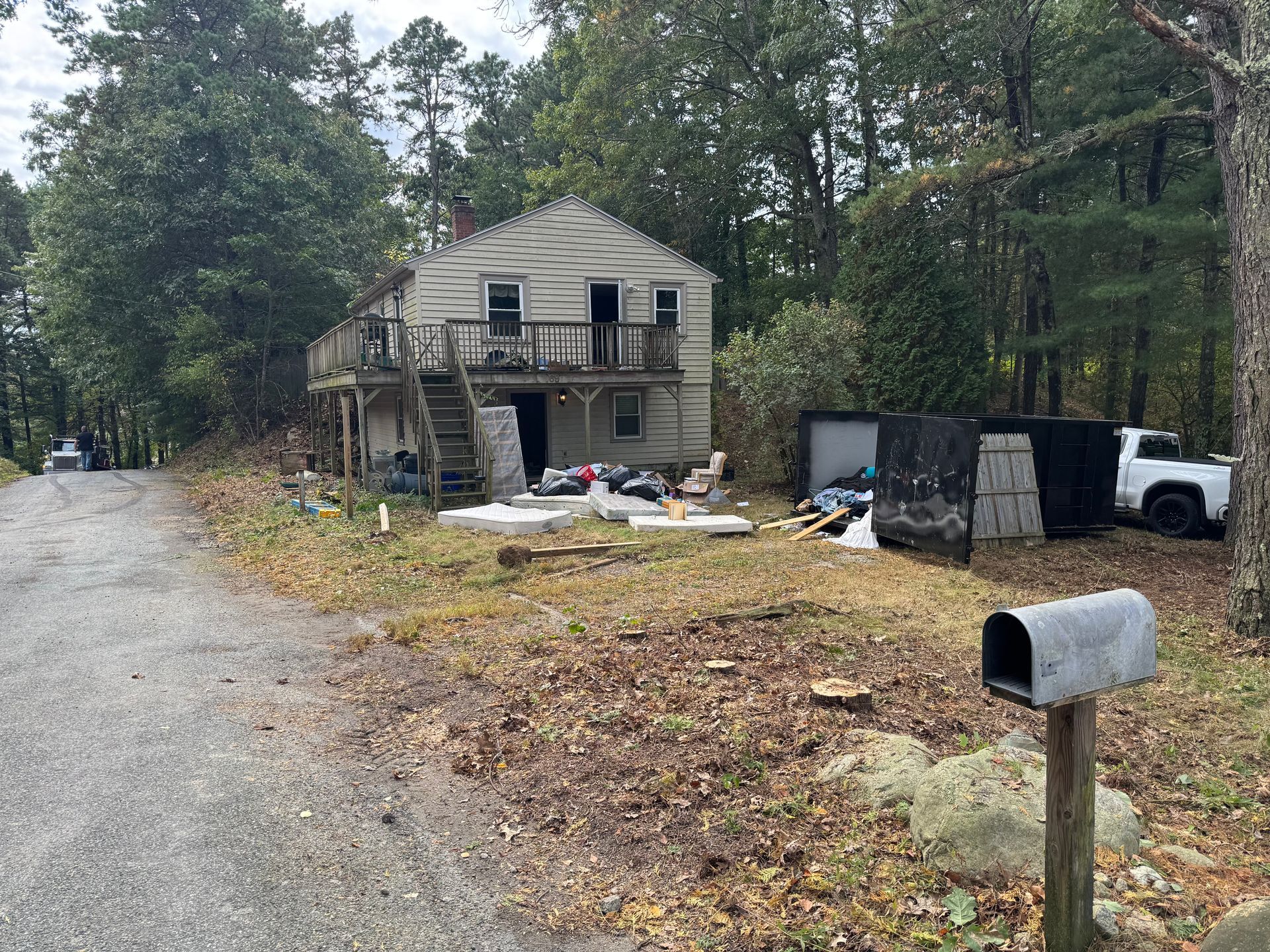 A weathered, two-story house with a wooden deck stands in a wooded area, with a dumpster and white truck nearby.