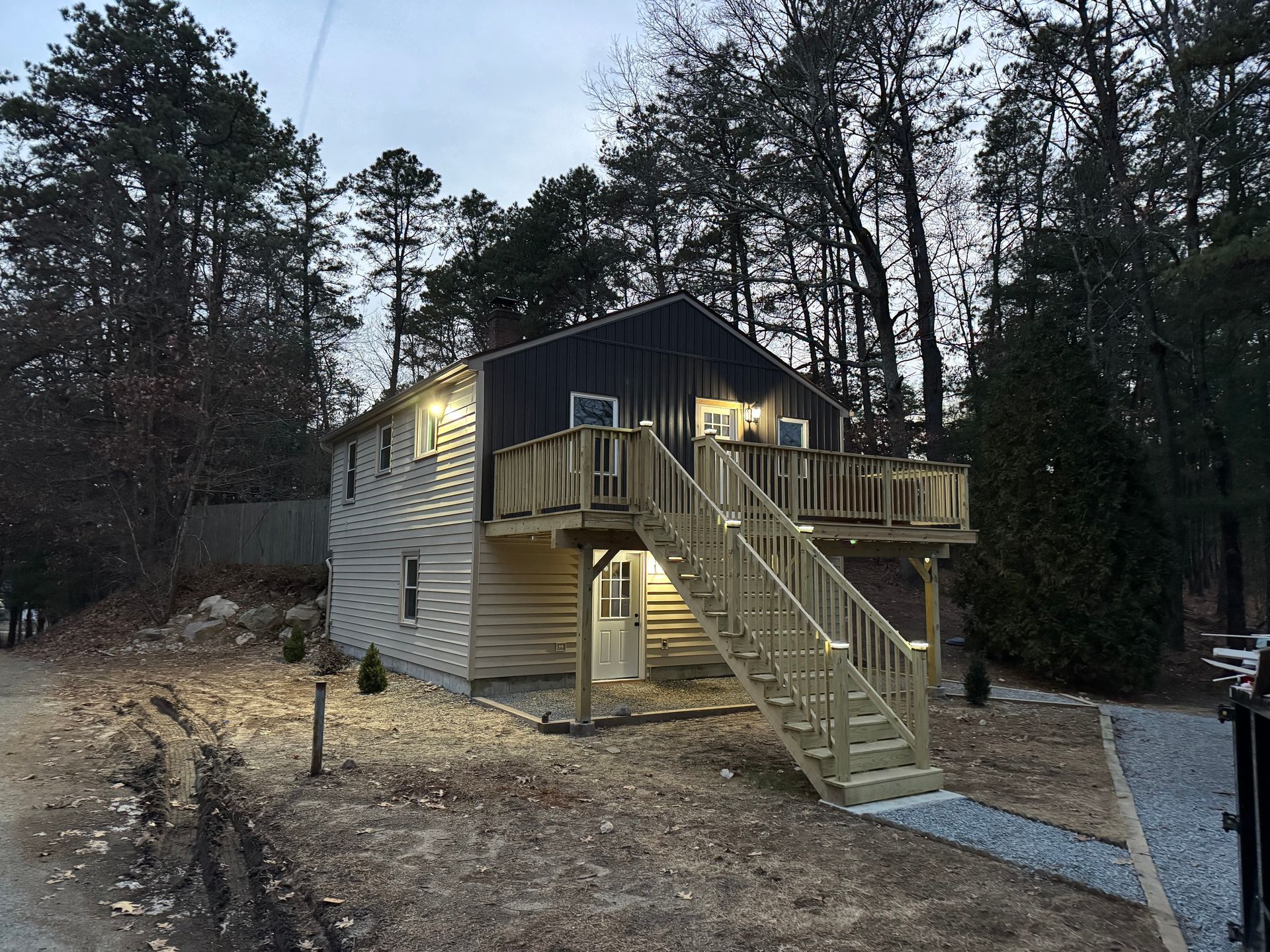 A two-story house with beige siding and a dark upper level, featuring an elevated wooden deck and staircase at dusk.