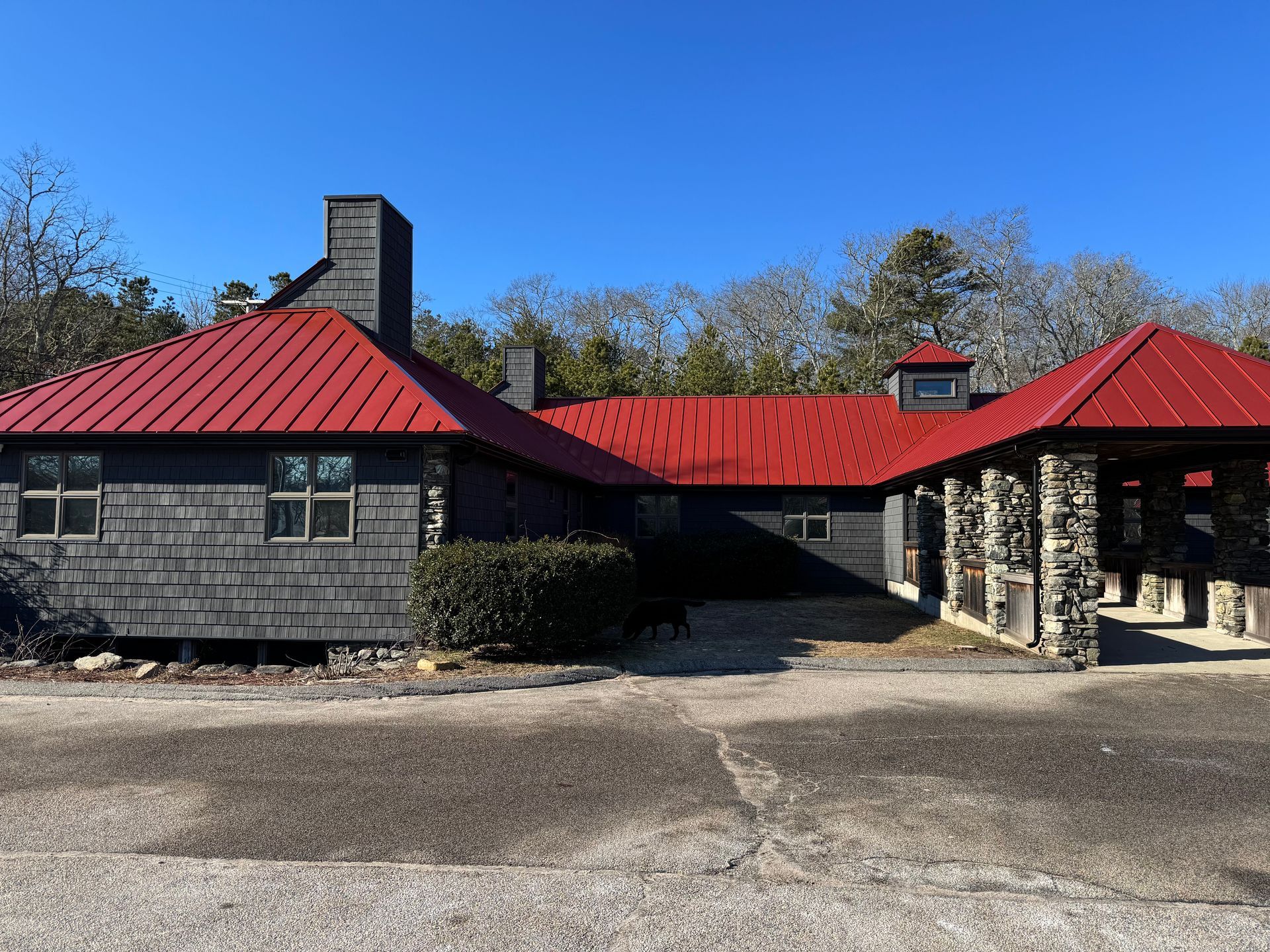 Single-story building with dark wood siding, a bright red metal roof, a stone-pillared porch, and a clear blue sky.