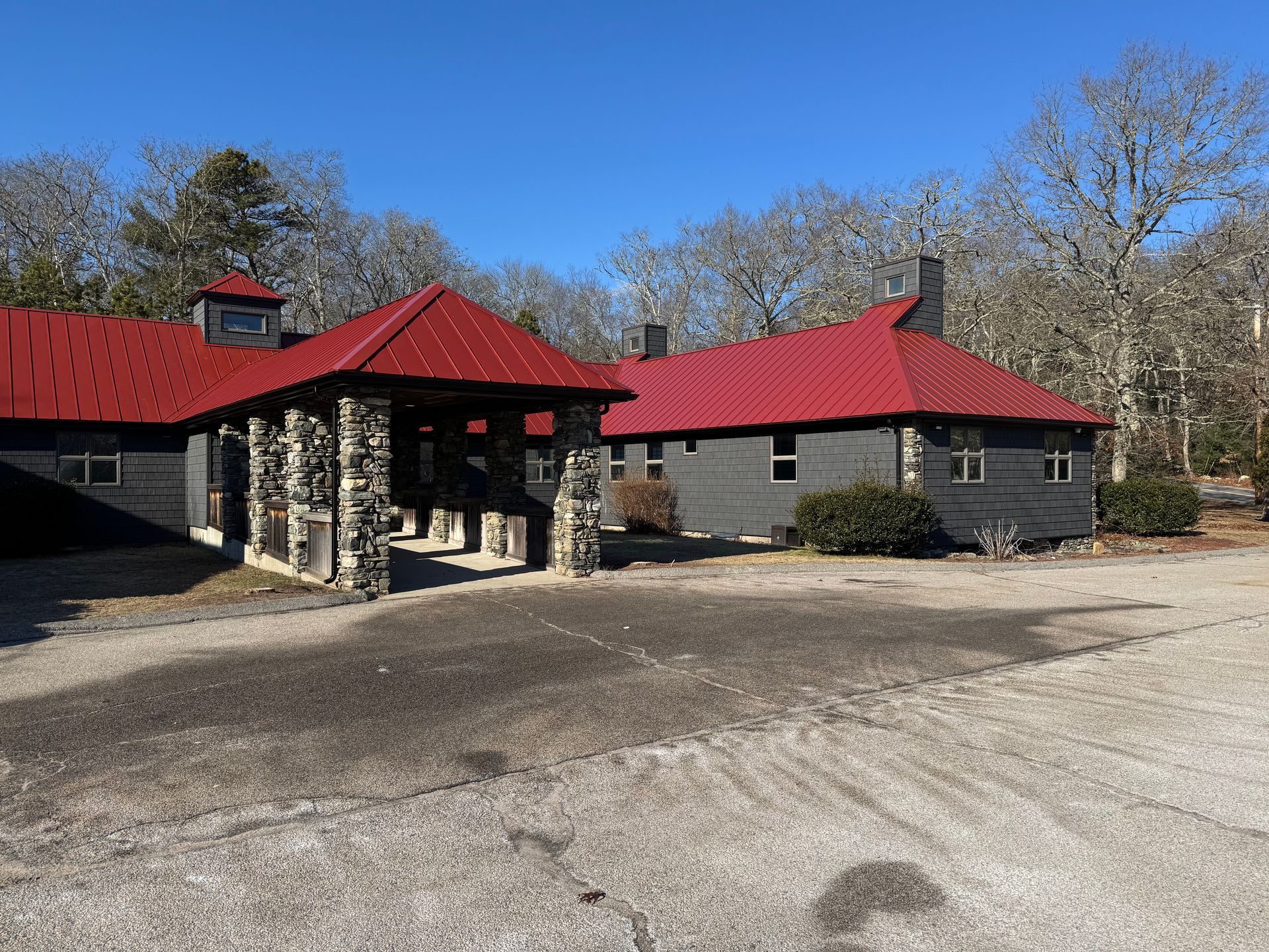 A dark gray building with a vibrant red metal roof and a stone-pillared entrance sits in a gravel lot surrounded by trees.