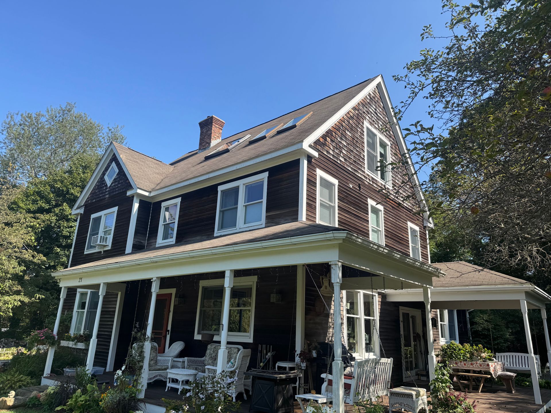 A two-story dark shingled house with a white wrap-around porch and peaked gables under a clear blue sky.