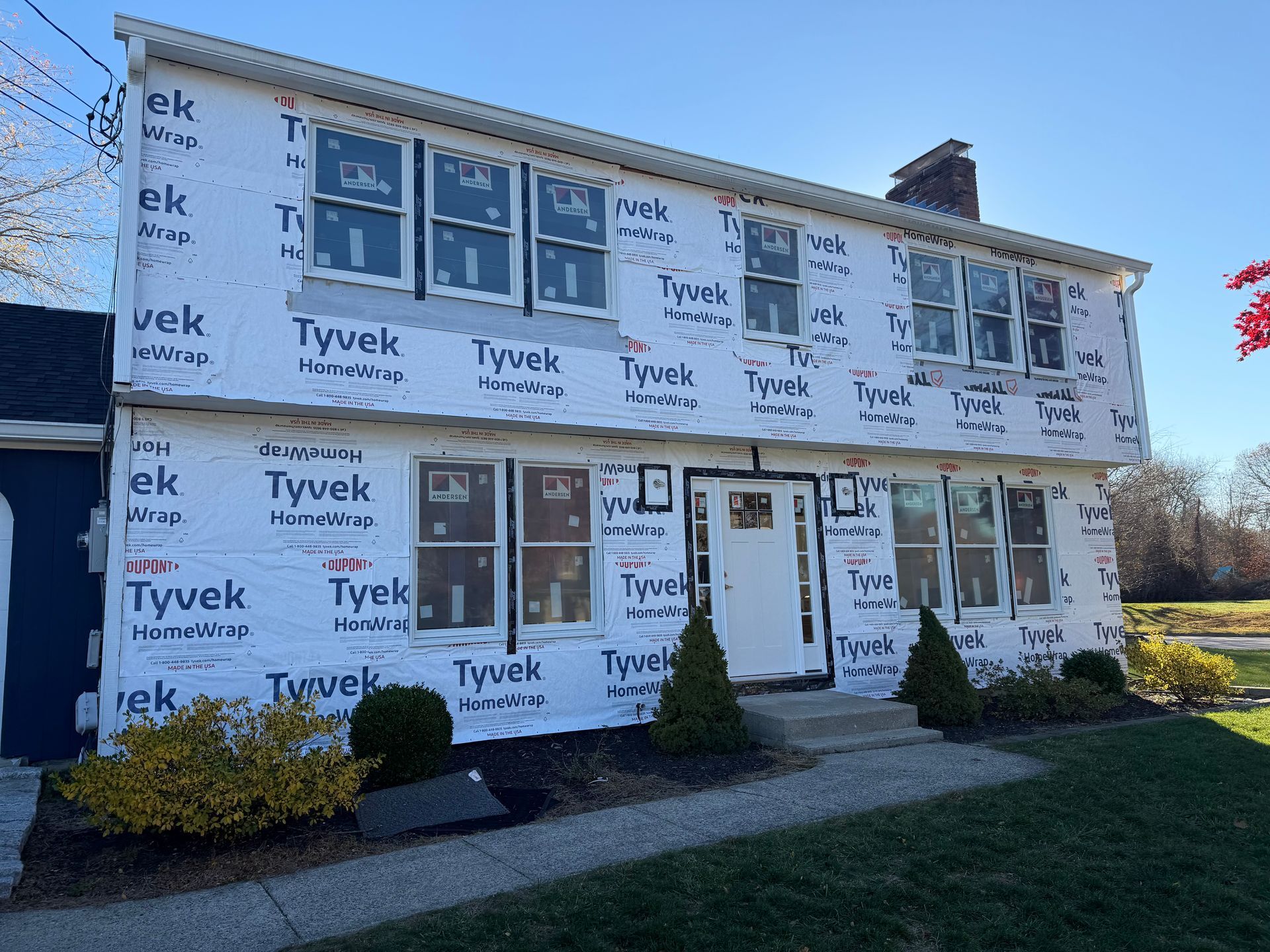 A two-story house under renovation, covered in white Tyvek weather-resistant barrier, viewed from the front yard.
