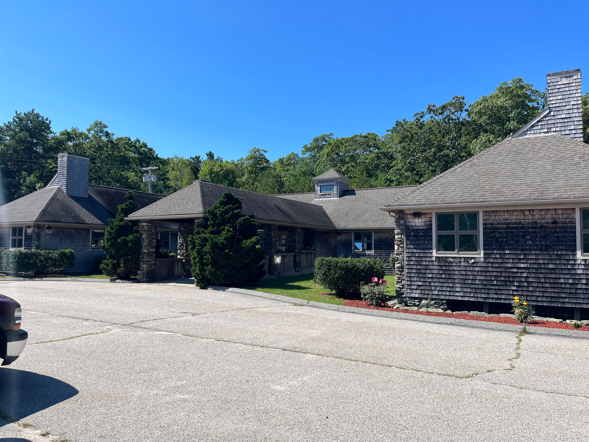 A row of weathered cedar-shingled buildings with stone chimneys, surrounded by trees under a clear blue sky.