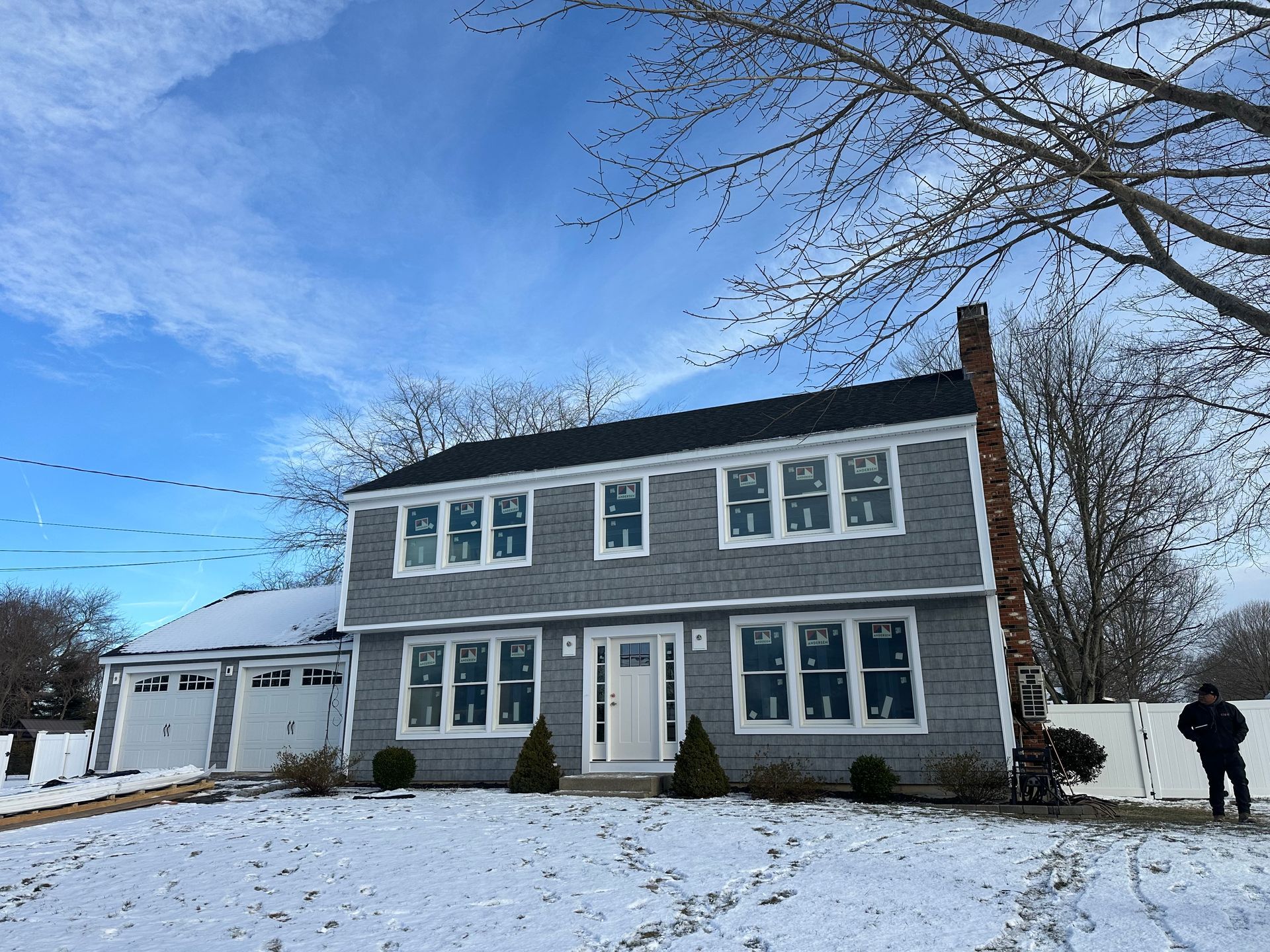 A two-story grey house with a white front door and garage, surrounded by snow under a bright blue sky.