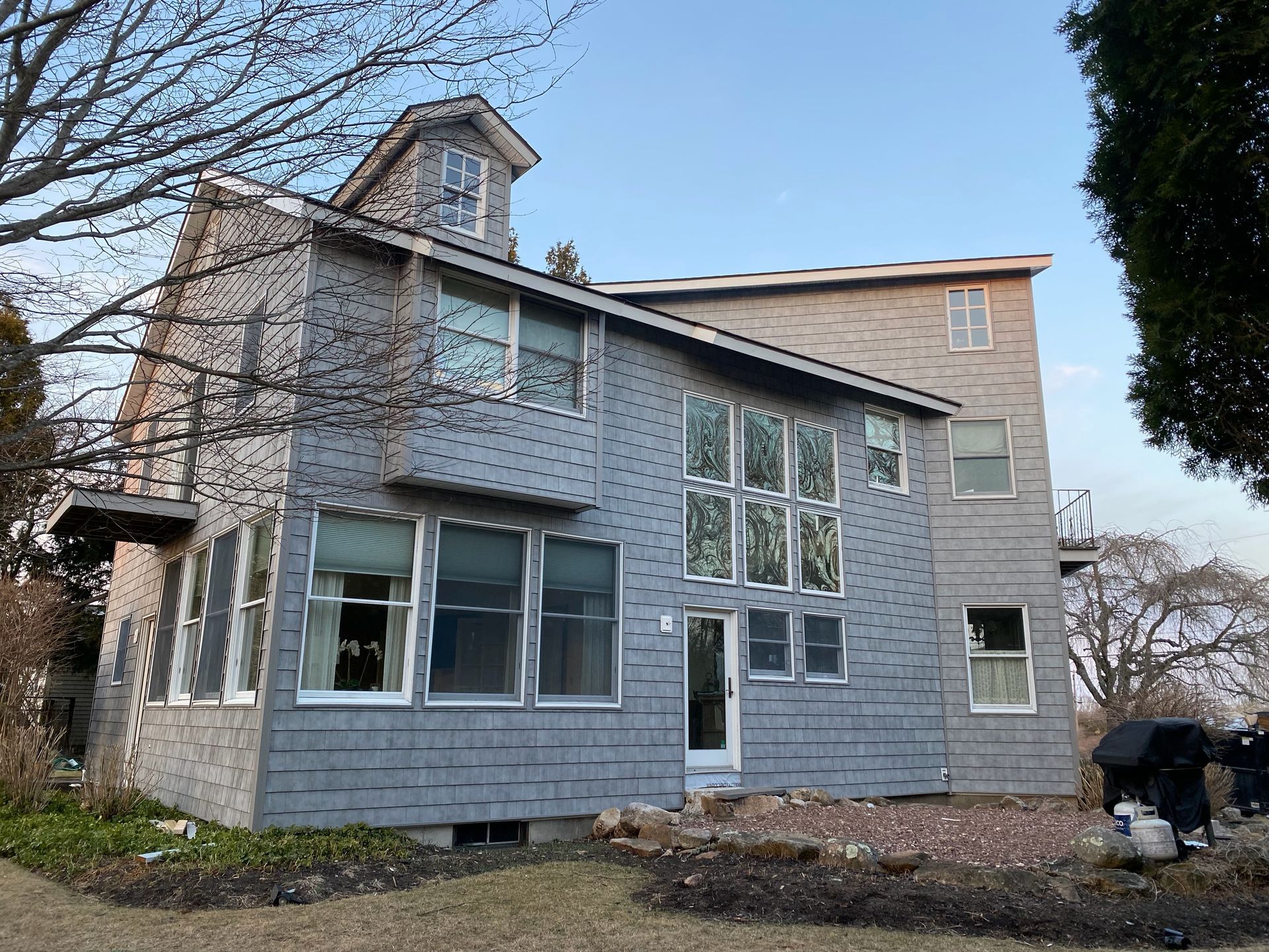 A two-story grey shingled house with an asymmetrical roof, large windows, and a partially graveled yard under a clear sky.