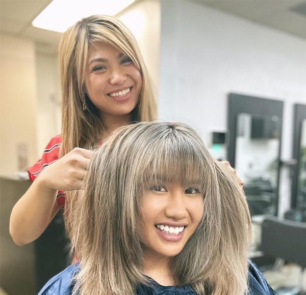 Woman getting hair styled in a salon; hairstylist with blonde hair smiles while adjusting client's cut.