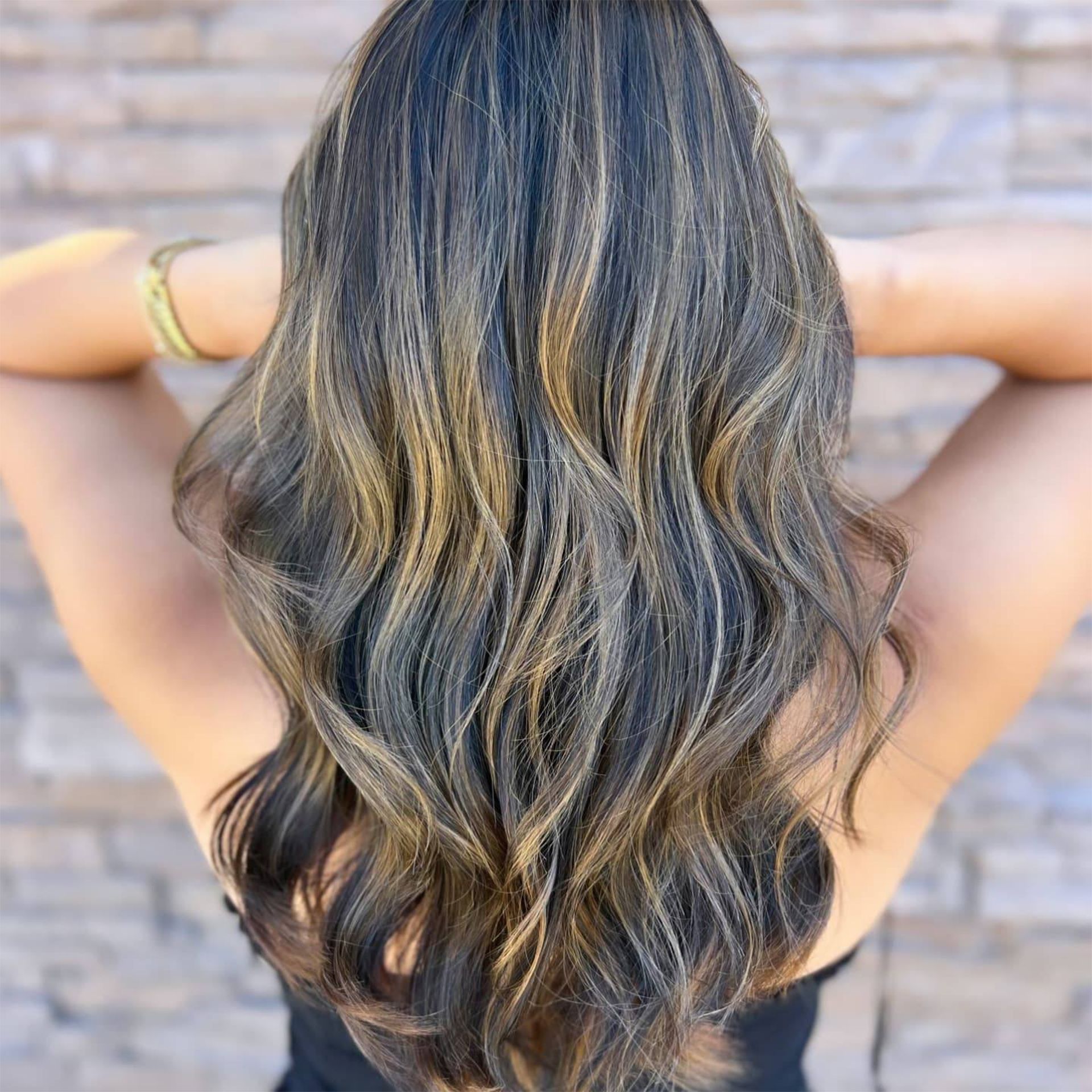 Back of a person with long, wavy hair, dark with blonde highlights, arms raised.